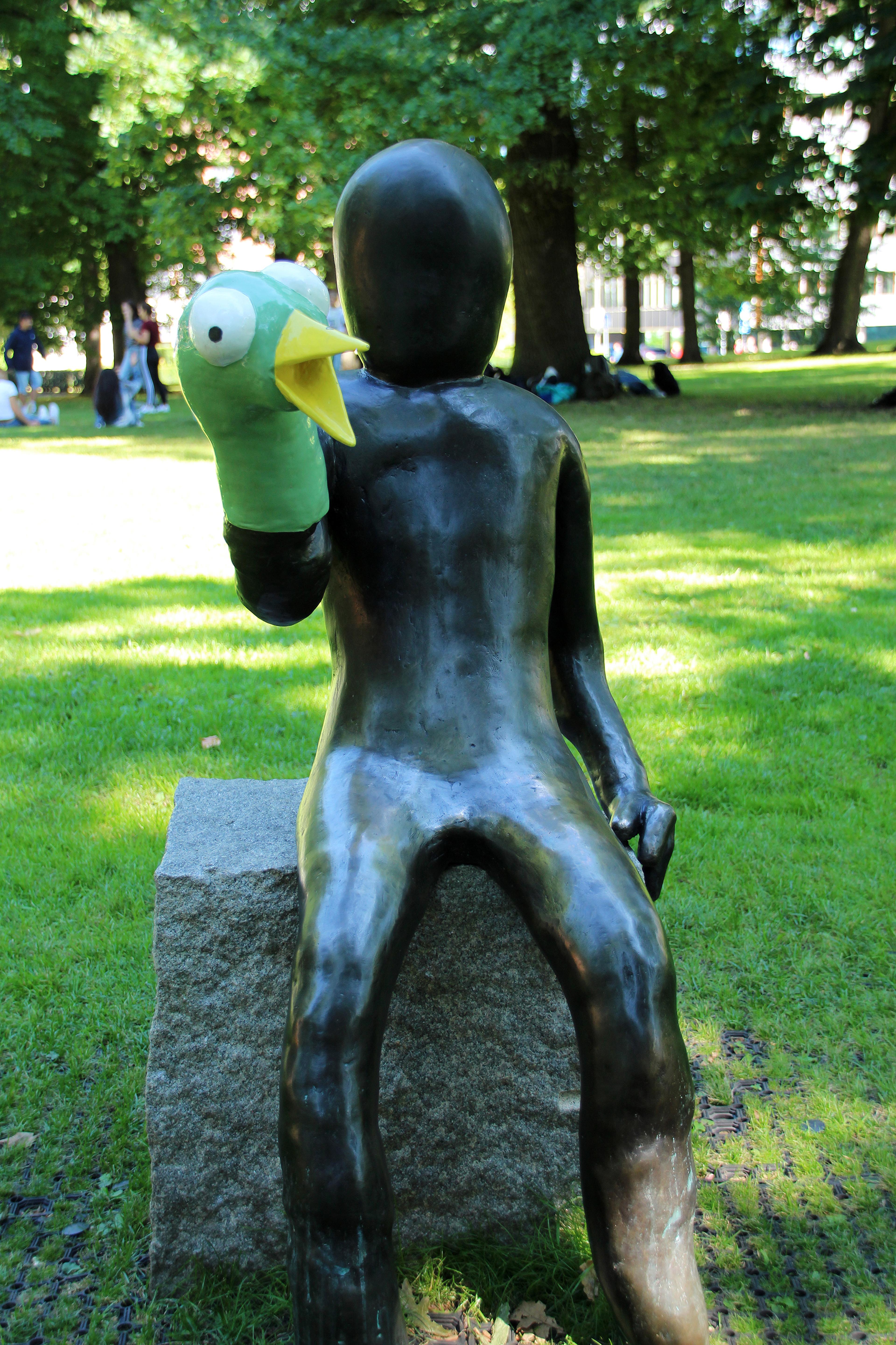 En støpt menneskefigur sitter på en stenblokk. Hodet er uten ansiktstrekk. Et grønt fuglehode med store øyne og åpent, gult nebb er tredd nedover den ene hånden som en dukke.