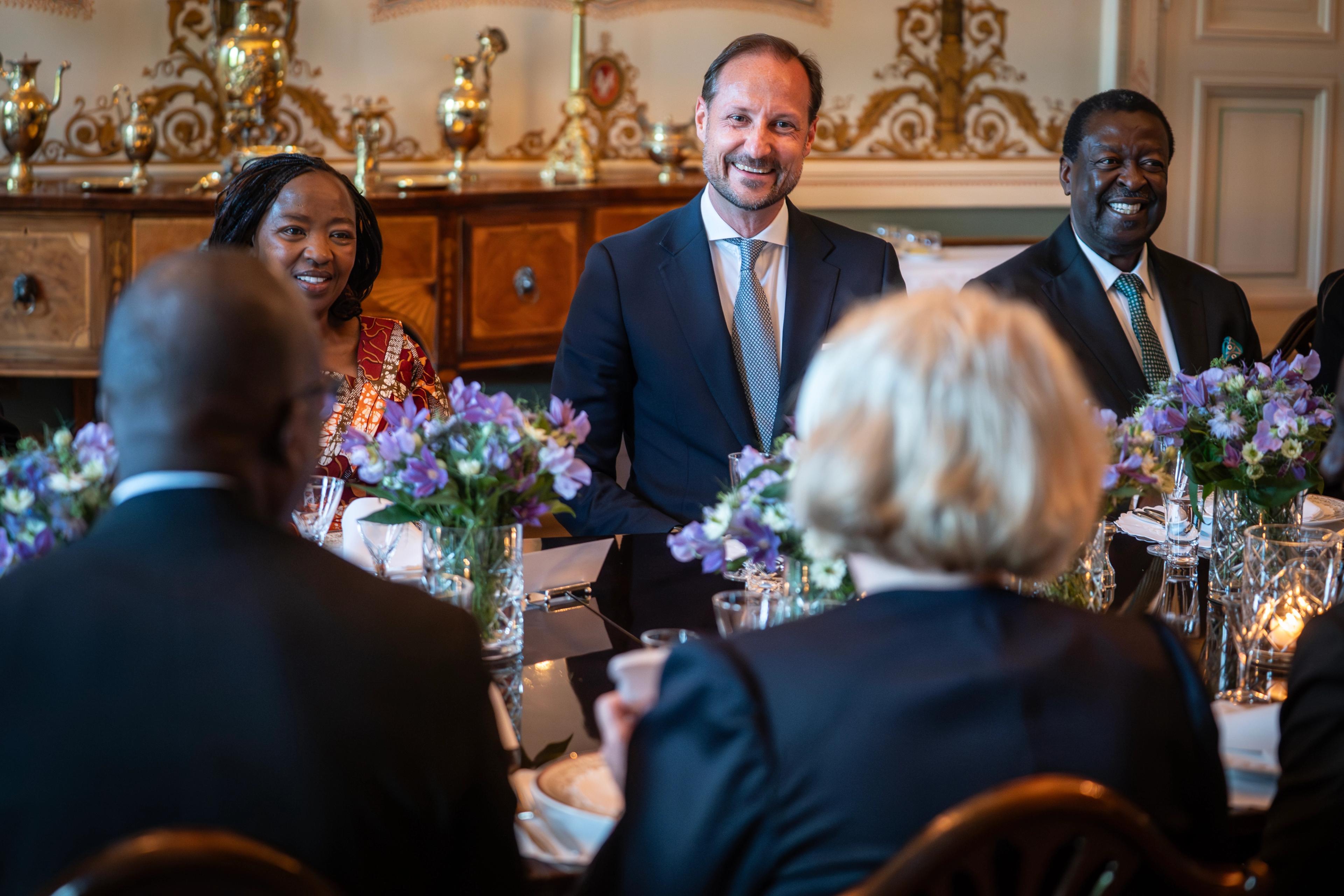 Kronprins Haakon og Rachel Ruto sitter ved eit bord og smiler til dei som sit med ryggen til.