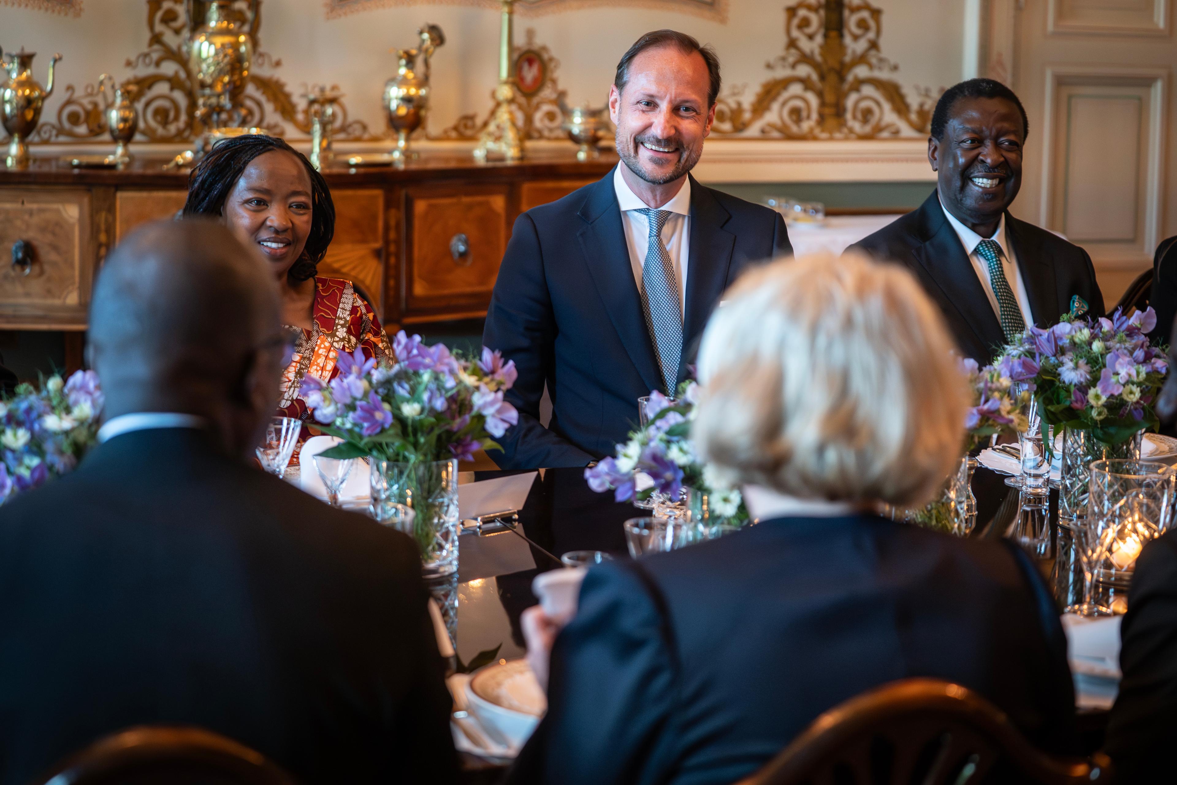 Kronprins Haakon og Rachel Ruto sitter ved eit bord og smiler til dei som sit med ryggen til.