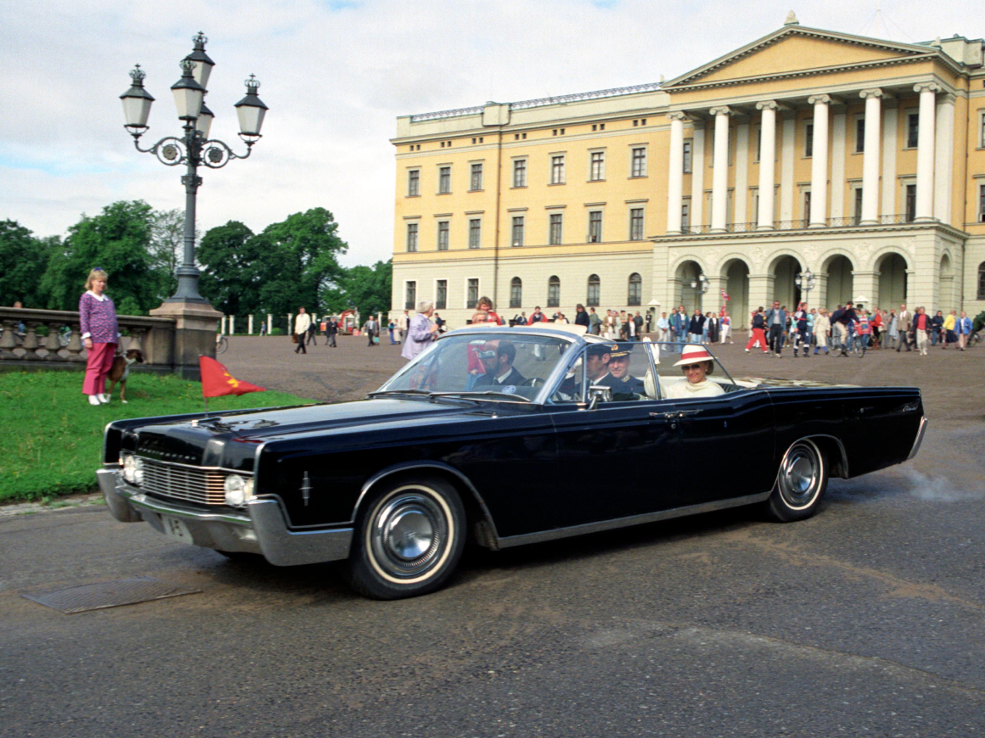 Veteranbilen A-5 på vei ned over Slottsplassen. Taket er nede, og Kongeparet vinker. Slottets fasade og mennesker på plassen i bakgrunnen.
