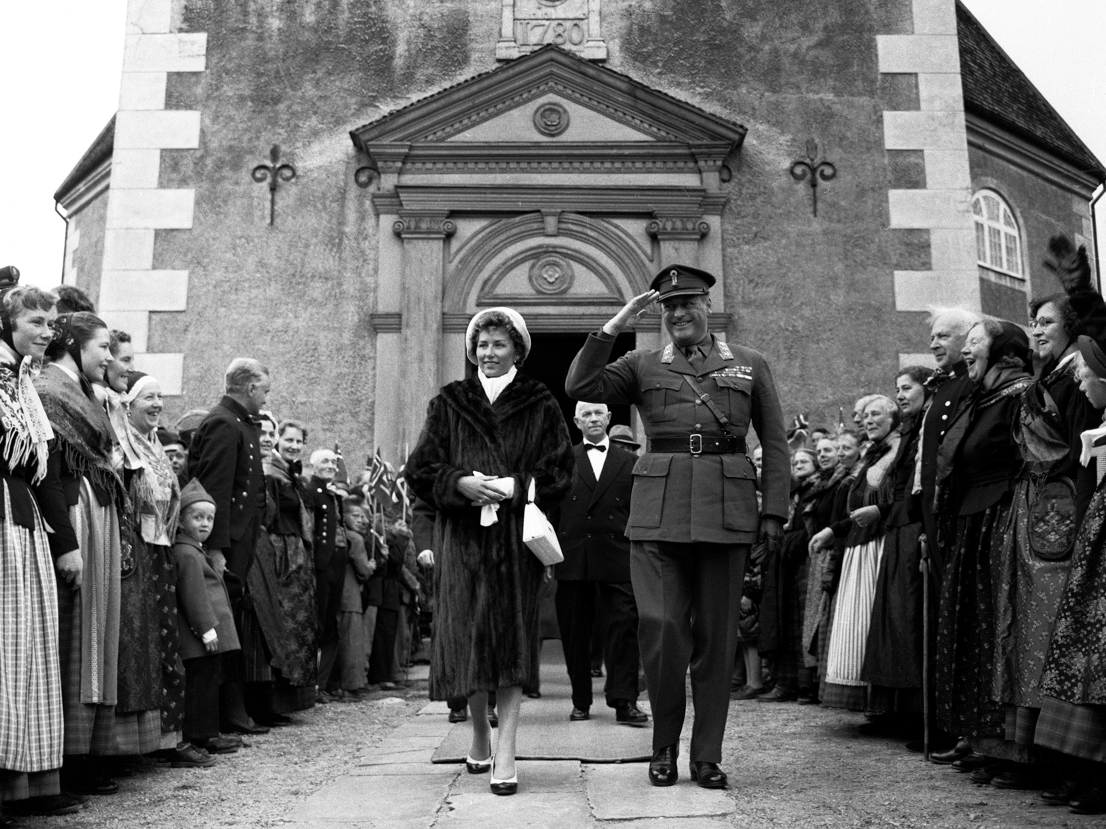 Sort-hvittbilde av Kongen og Prinsessen på vei ned bakken utenfor Røros kirke. Kirkedøren er bak dem, og stien er omkranset av mennesker.