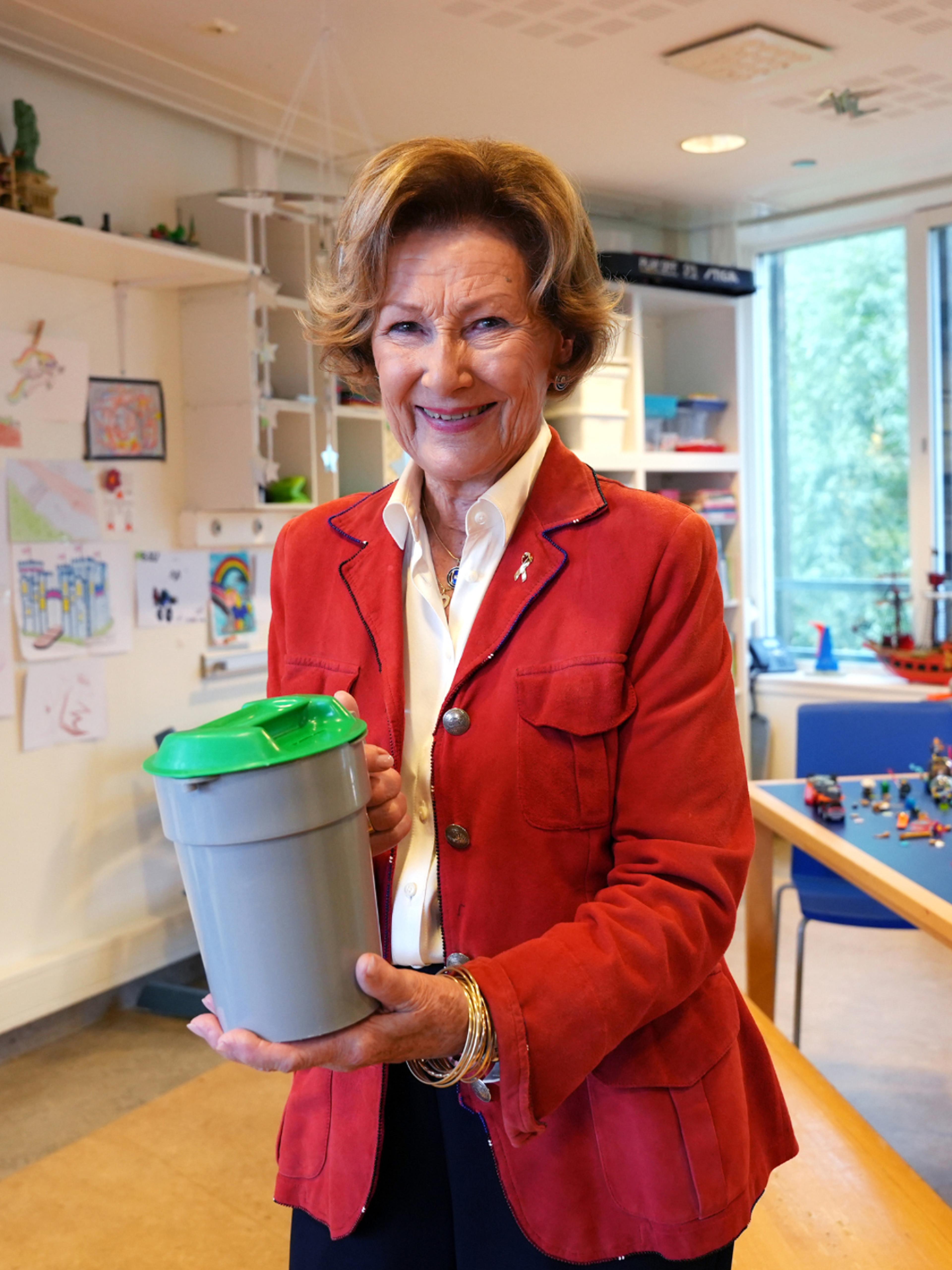 En smilende dronning i rød jakke holder fram en innsamlingsbøsse fra TV-aksjonen.