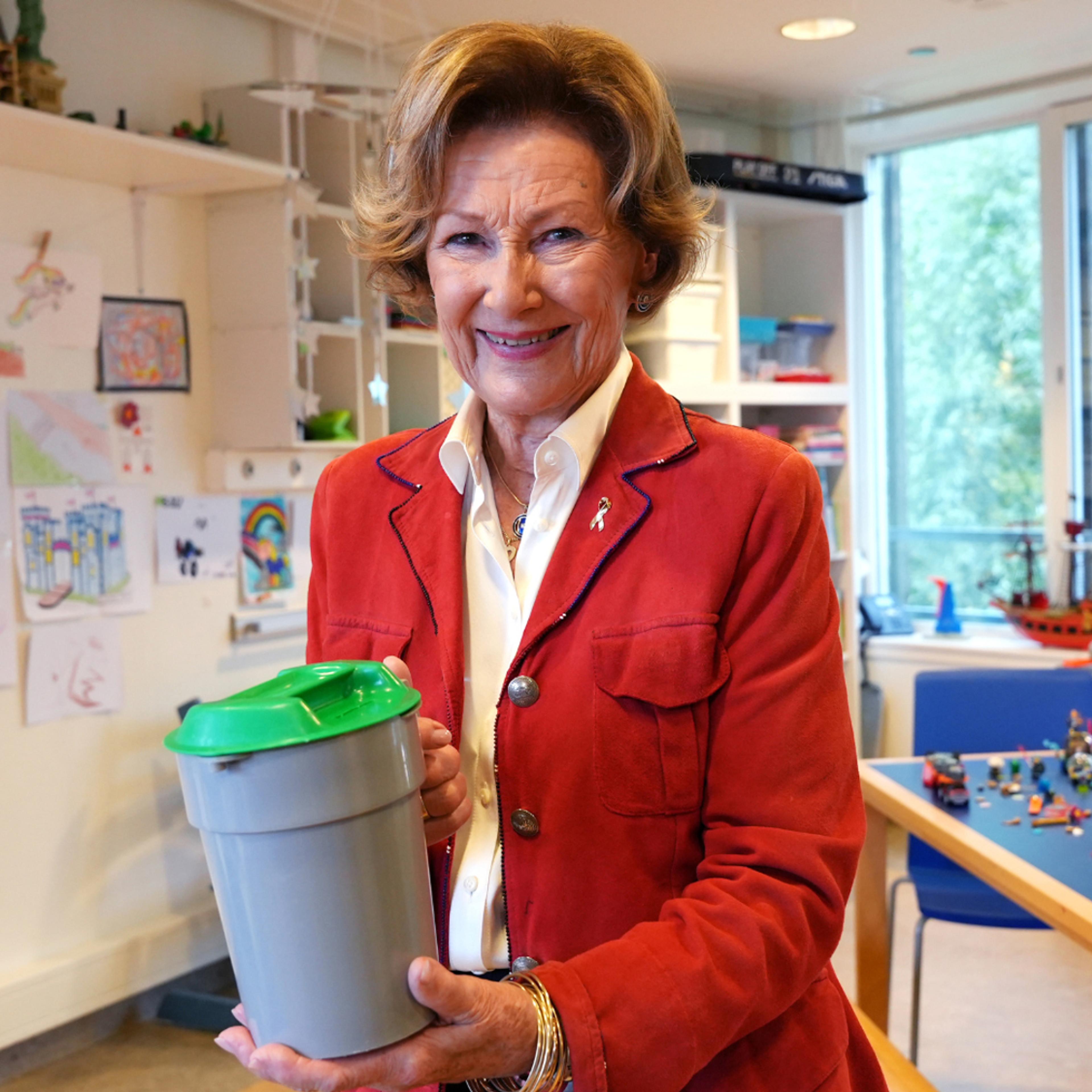 En smilende dronning i rød jakke holder fram en innsamlingsbøsse fra TV-aksjonen.