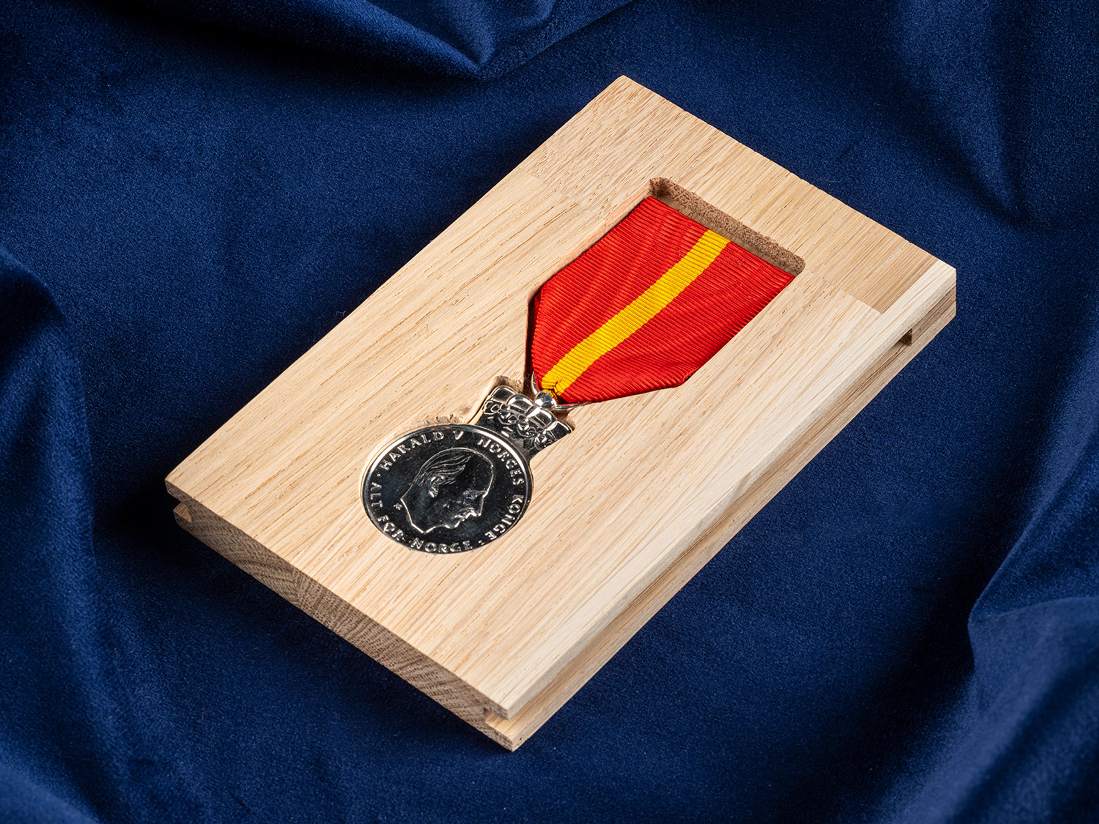 Medalje i sølv, med en rødt bånd med gul stripe, ligger i en formlaget eske av eik. Esken ligger på et dypblått klede.