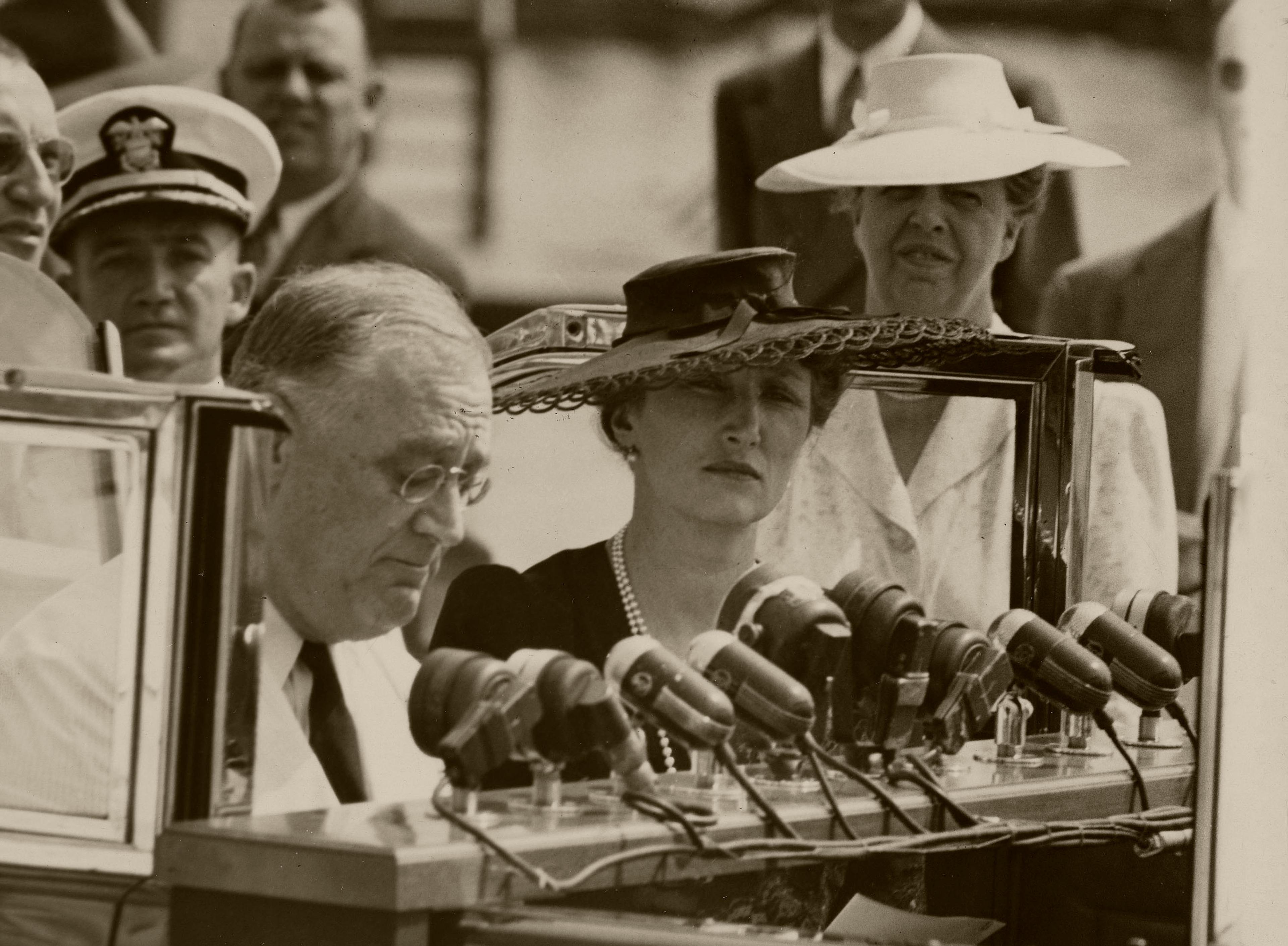Kronprinsesse Märtha står ved siden av Presidenten, mens han taler foran et stort antall mikrofoner.