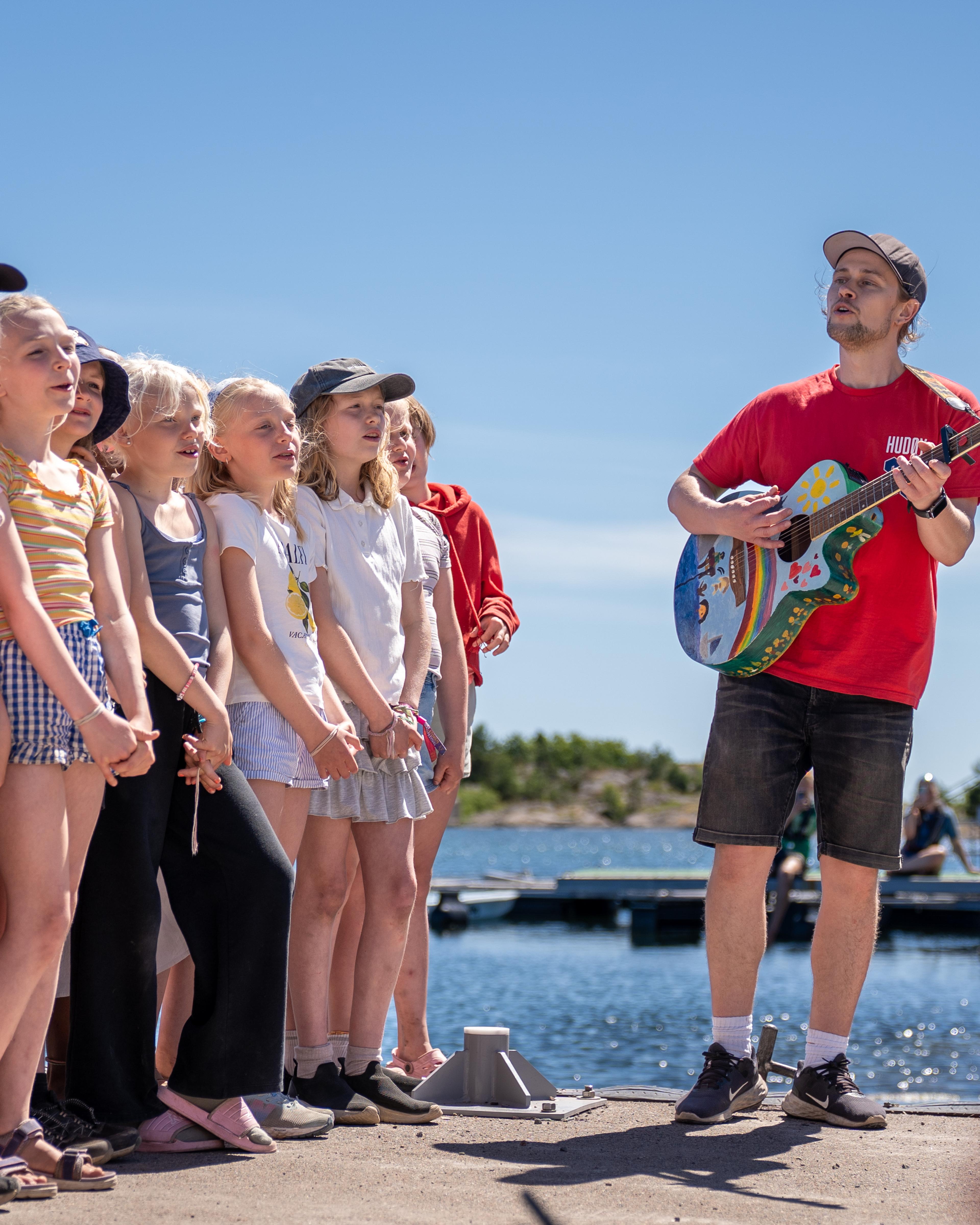 Christopher står med gitar. Jenter står på rekke og synger.