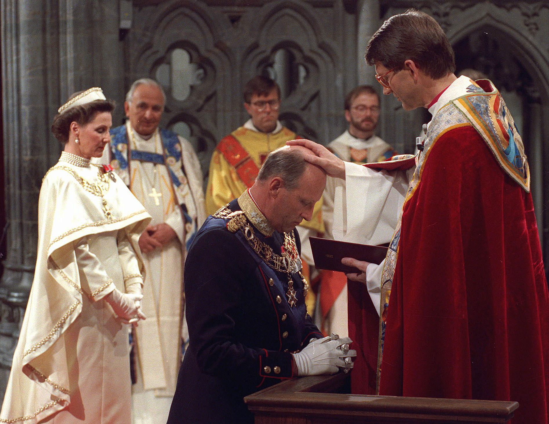 Kong Harald kneler foran biskopen, som holder hånden på Kongens hode. Biskopen har rød kappe, Kongen er i gallauniform. Dronning Sonja står i bakgrunnen i sin hvite kroningskjole.