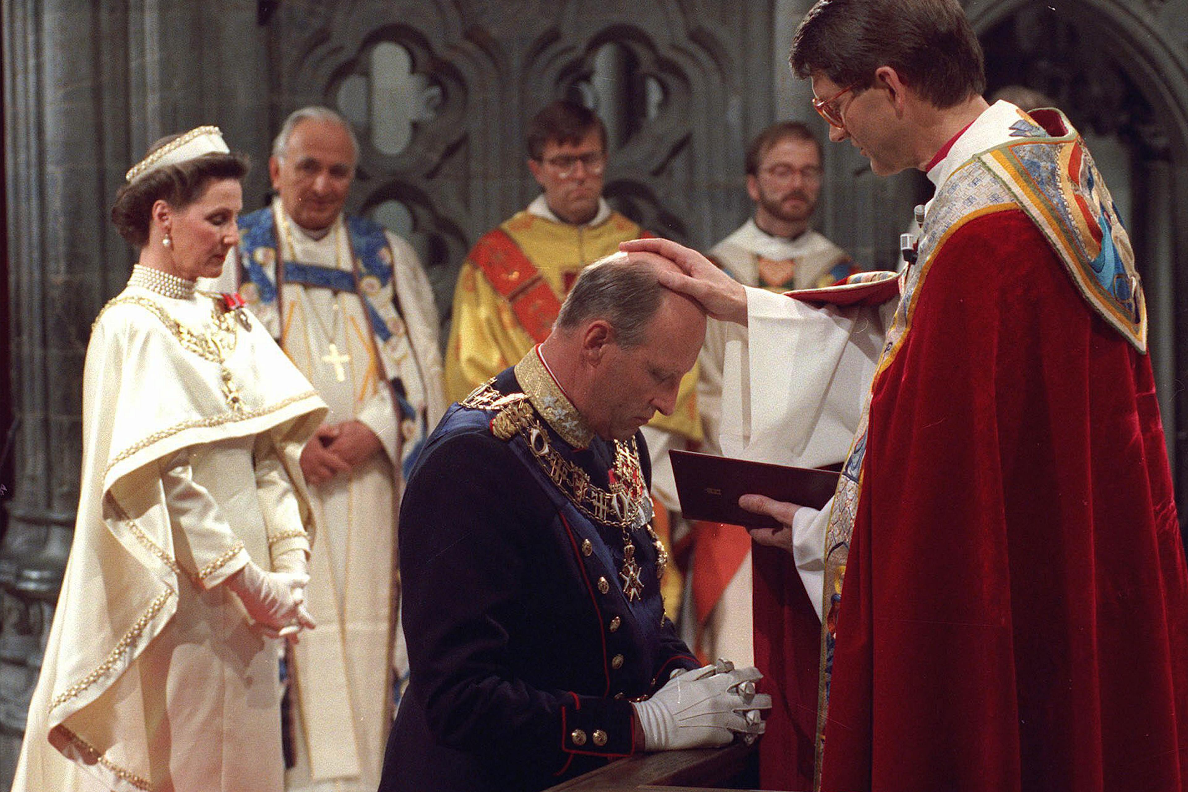 Kong Harald kneler foran biskopen, som holder hånden på Kongens hode. Biskopen har rød kappe, Kongen er i gallauniform. Dronning Sonja står i bakgrunnen i sin hvite kroningskjole.