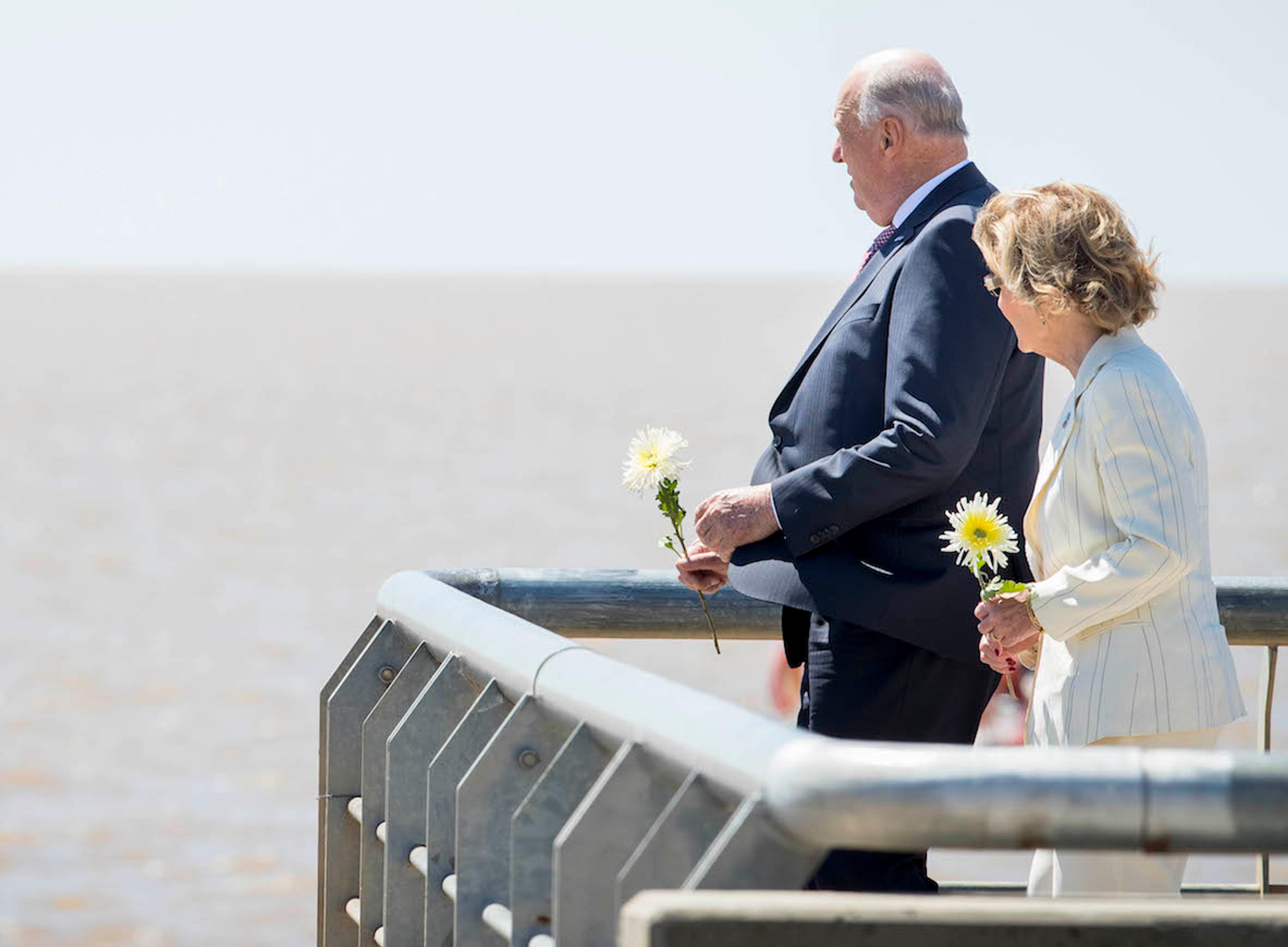 Kongen og Dronningen står med hvite roser i hendene og ser utover elven fra en utsiktsplattform.