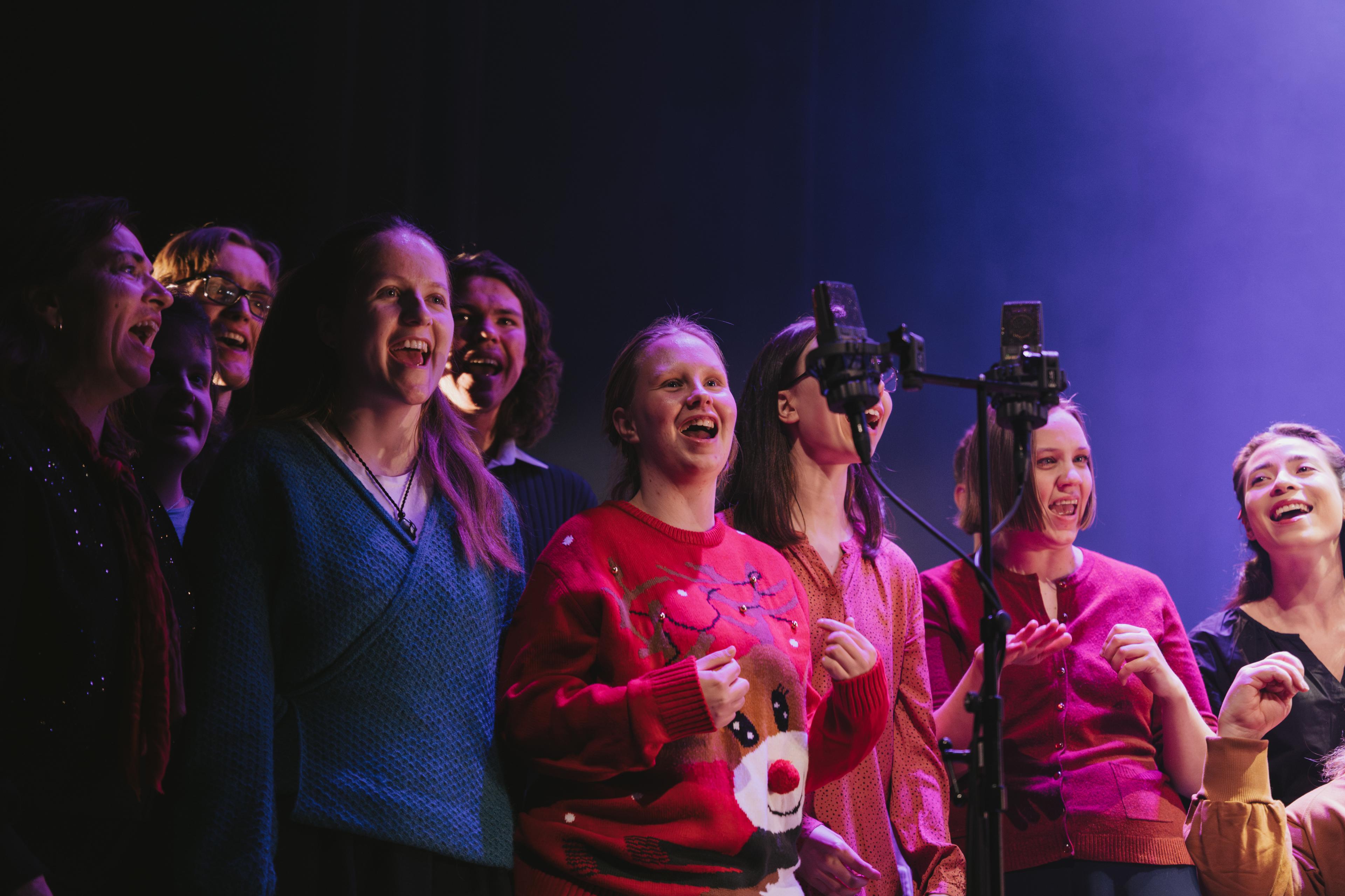 Eit kor på omlag 10 songarar syng inn i ein mikrofon.