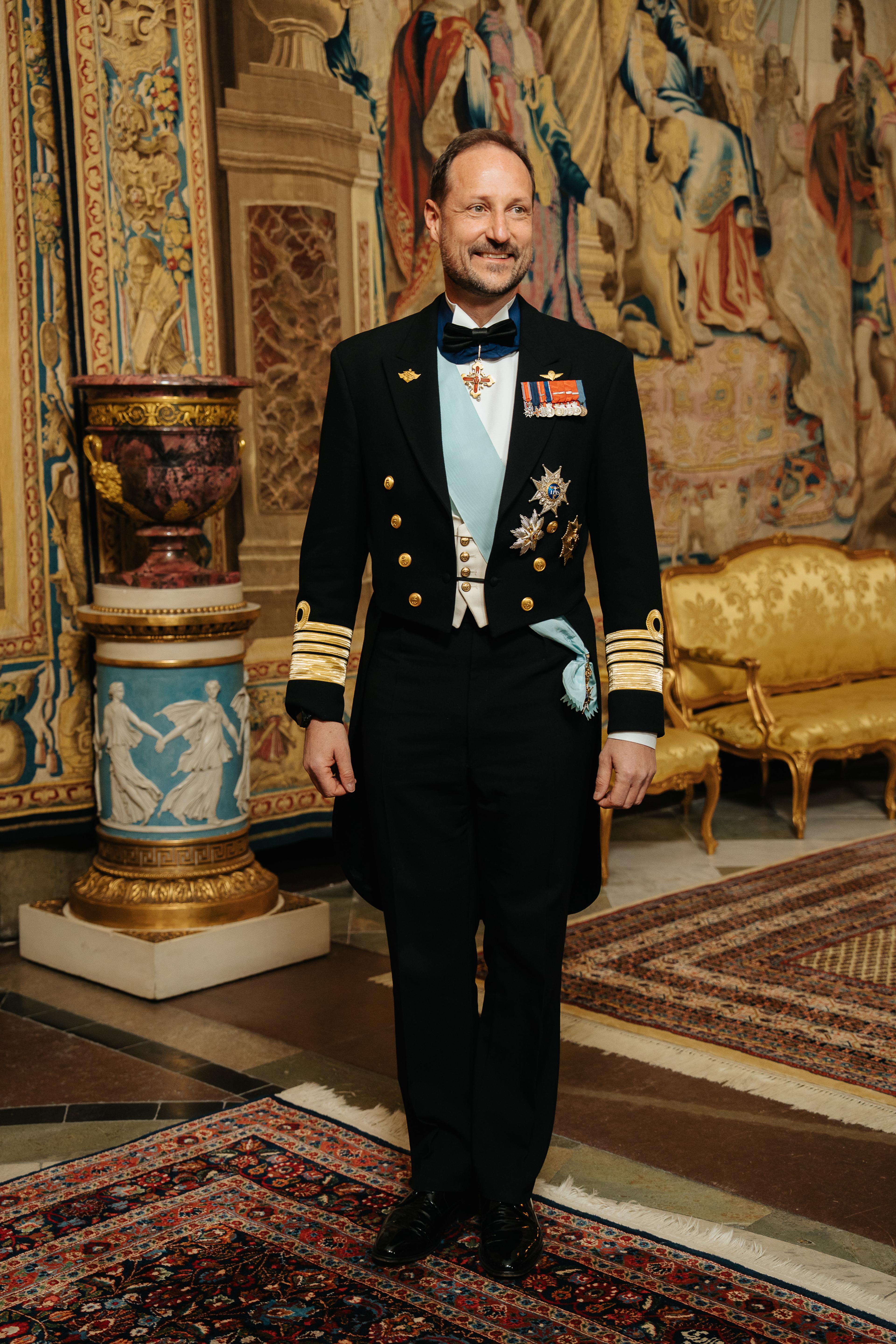 Kronprinsen kledd i marinens galla foran et stort veggteppe (gobelin).