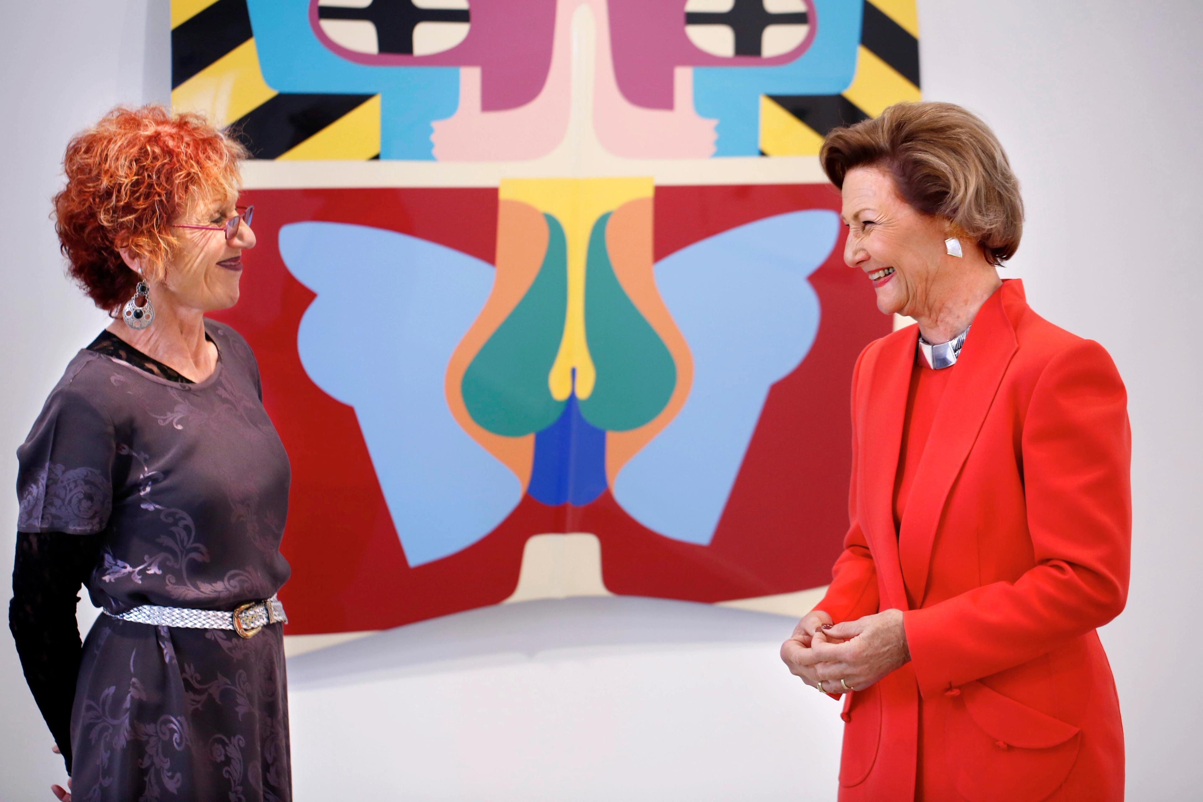 Judy Chicago og Dronning Sonja ser på hverandre og smiler. I bakgrunnen ser vi et stort verk av kunstneren på veggen.