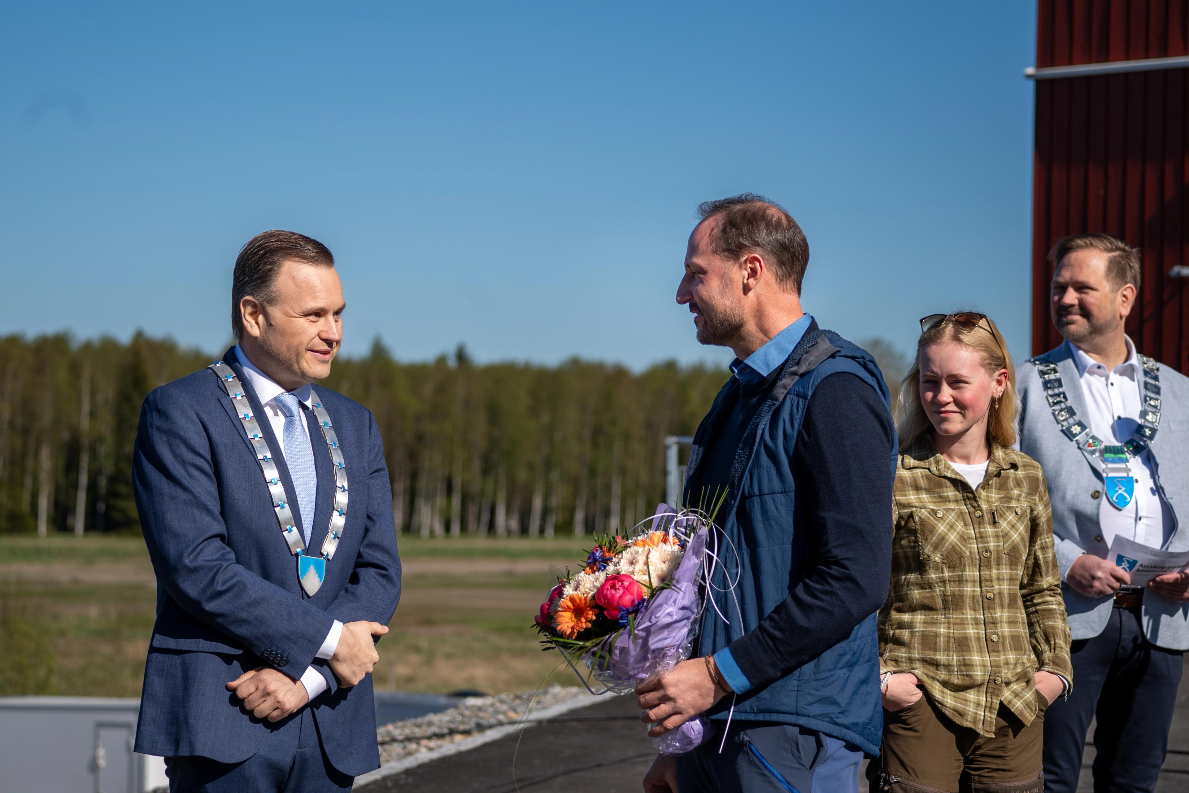 Kronprins Haakon står med blomstebukett i hånden, i samtale med fylkesordføreren med fylkeskjede rundt halsen.