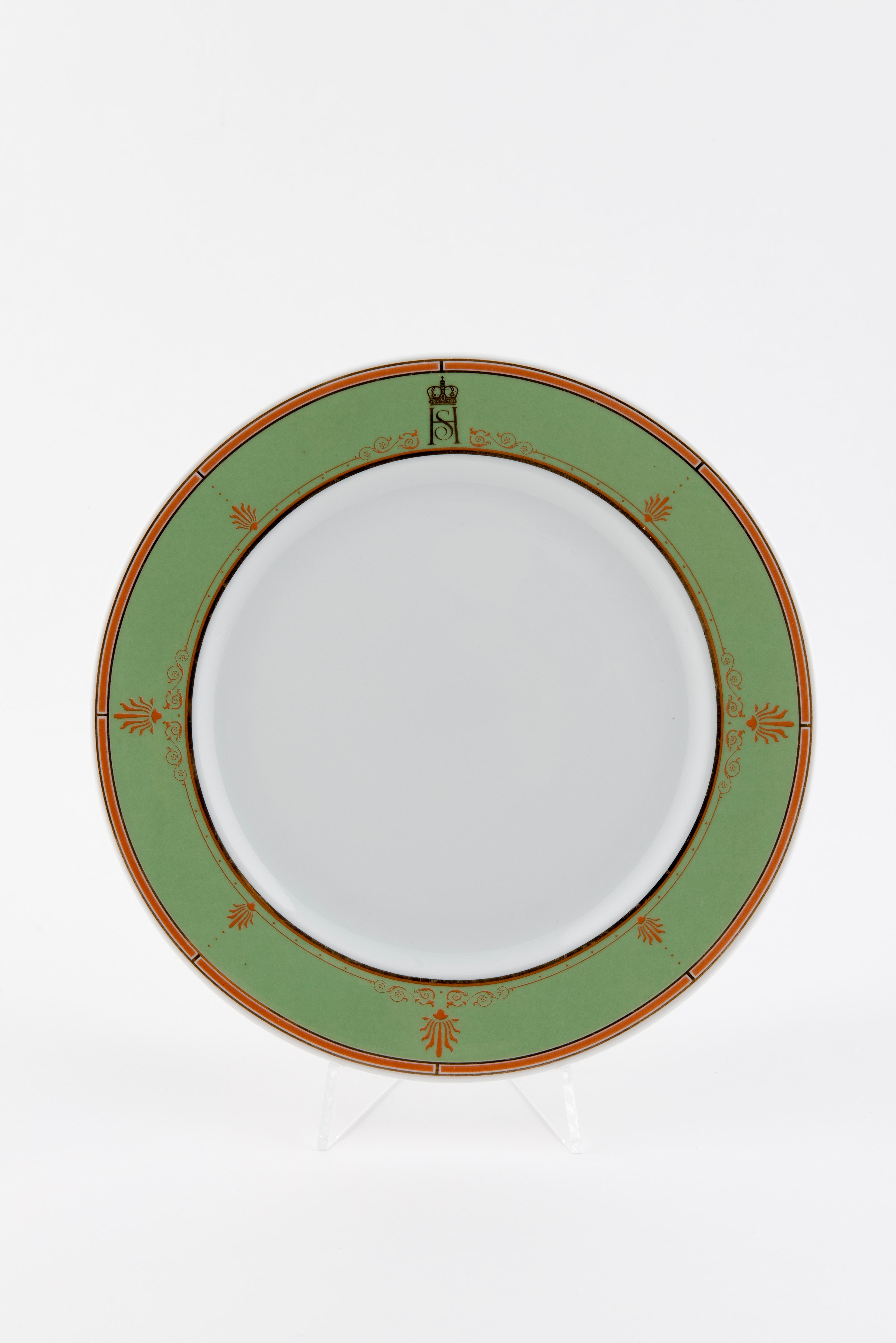 En hvit tallerken med en bred grønn kant med rød-orange dekor. En tynn stripe i samme rødfarge løper helt ytterst. Kongeaprets fellesmonogram står mot kanten øverst.
