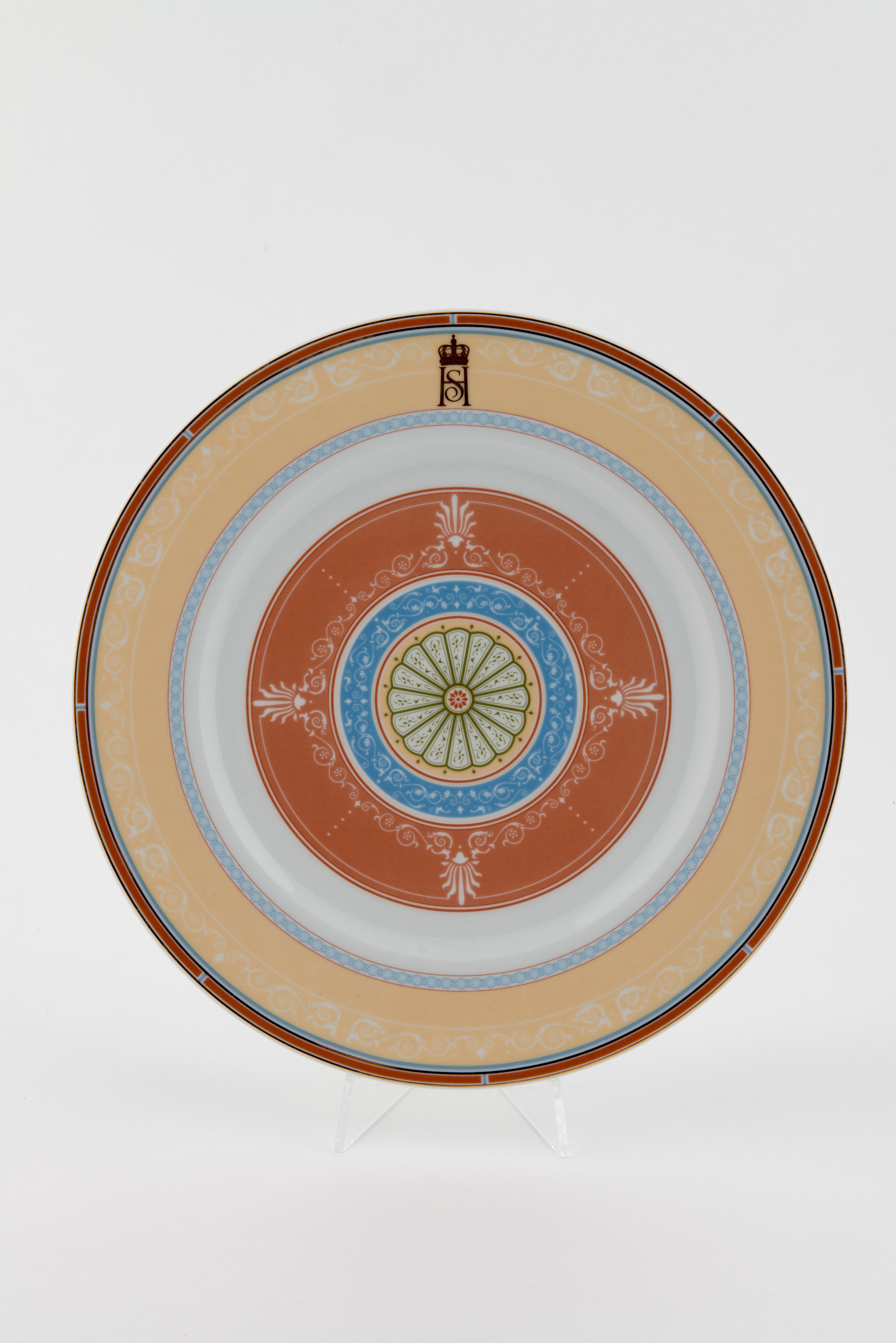 En tallerken med mønster inspirert av spisesalens dekor. Det er syv sirkler med ulik bredde og farge, med forsiktig dekor i de ulike feltene. Kongeparets fellesmonogram mot kanten øverst.