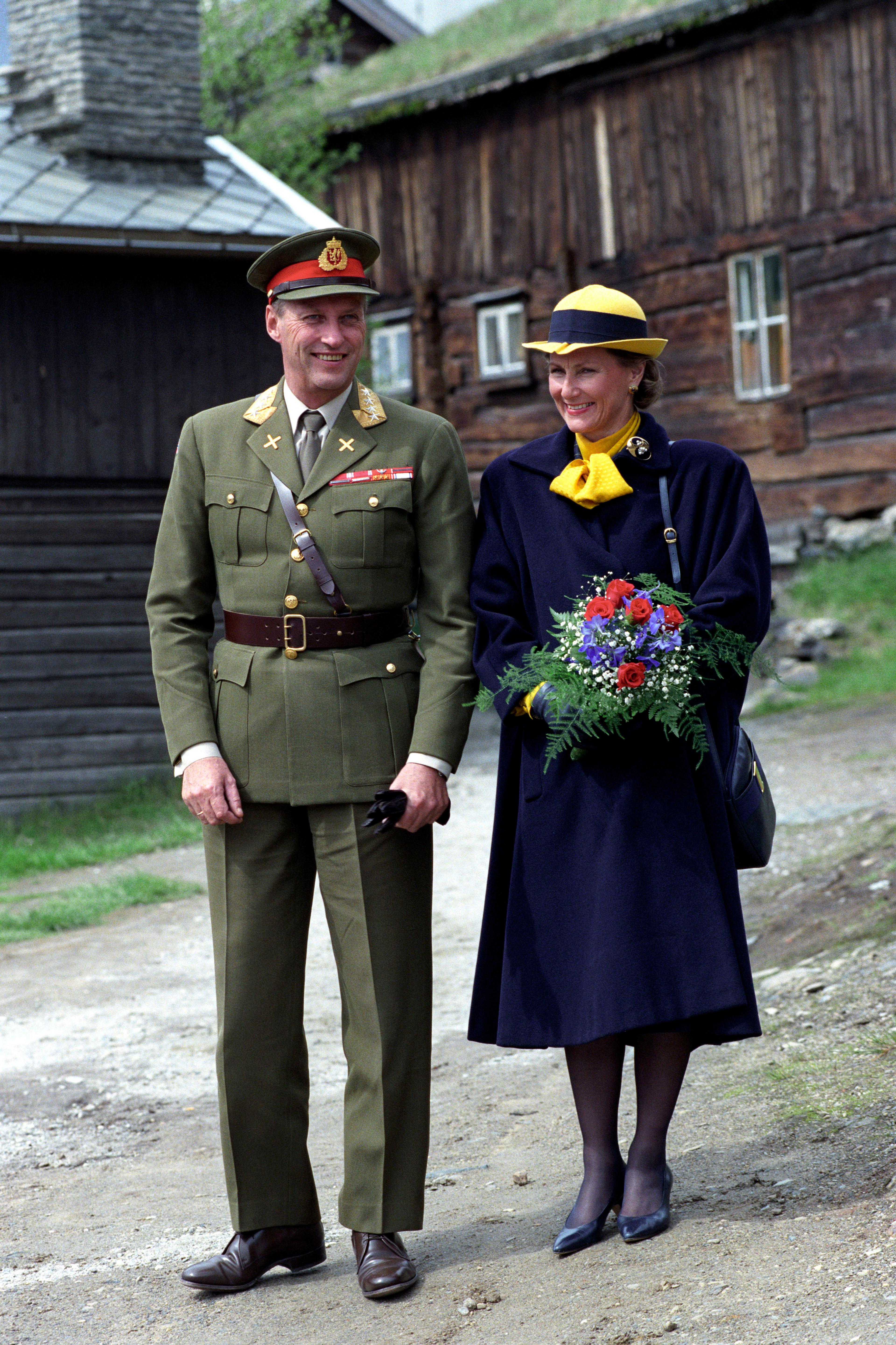 Et smilende kongepar med Røros' gamle trehusbebyggelse i bakgrunnen.