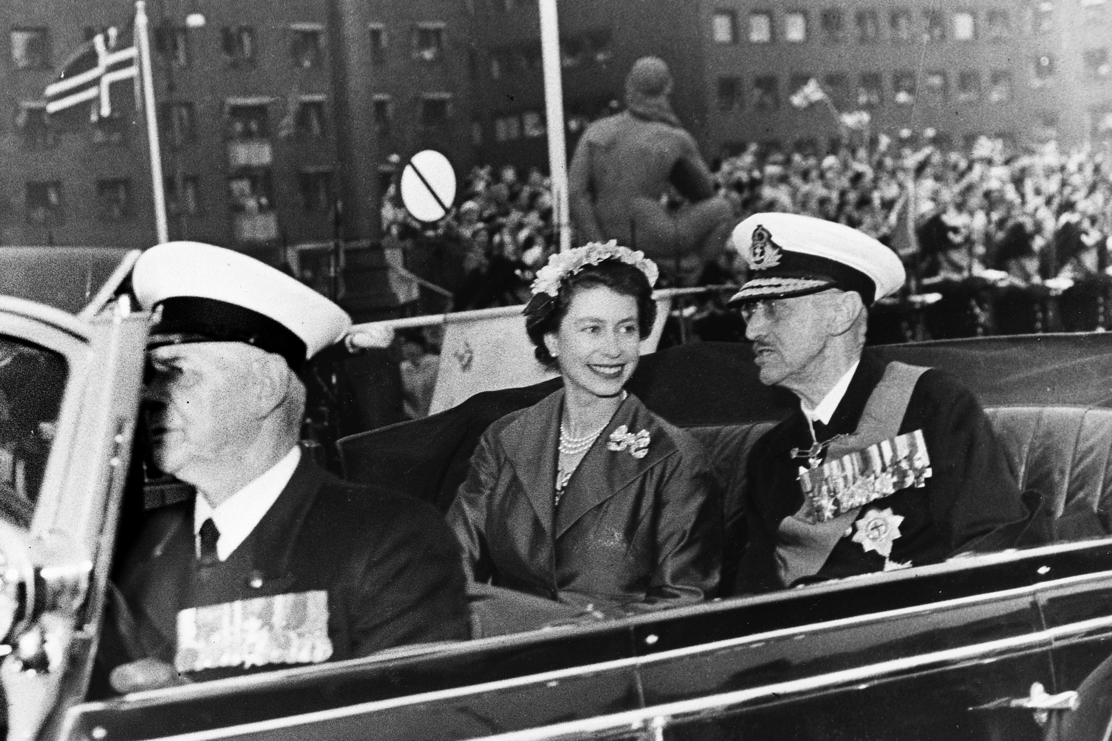 Sort-hvitt bilde av Kong Haakon og Dronning Elizabeth i åpen bil. Dronningen lytter til Kong Haakon med et stort smil.