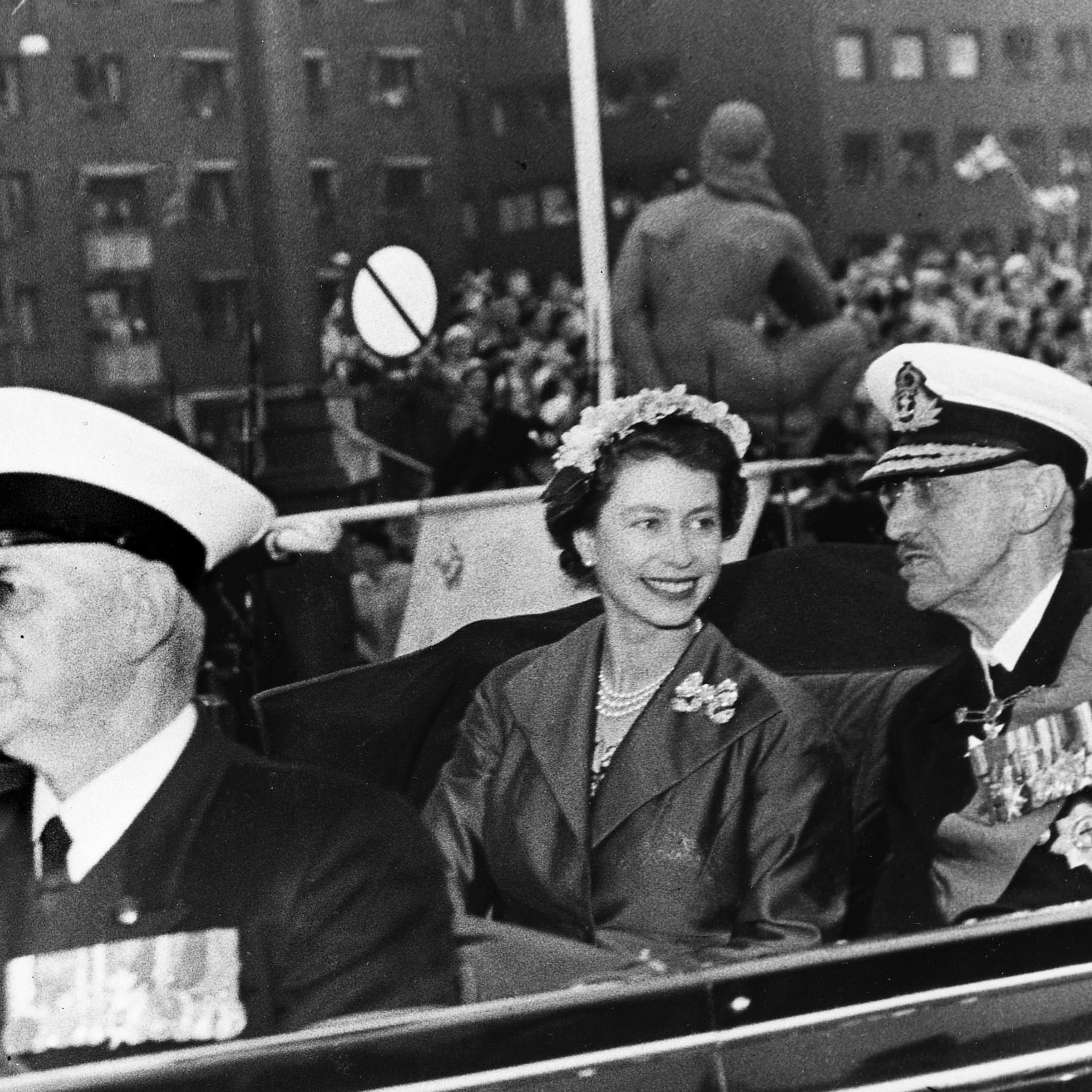 Sort-hvitt bilde av Kong Haakon og Dronning Elizabeth i åpen bil. Dronningen lytter til Kong Haakon med et stort smil.