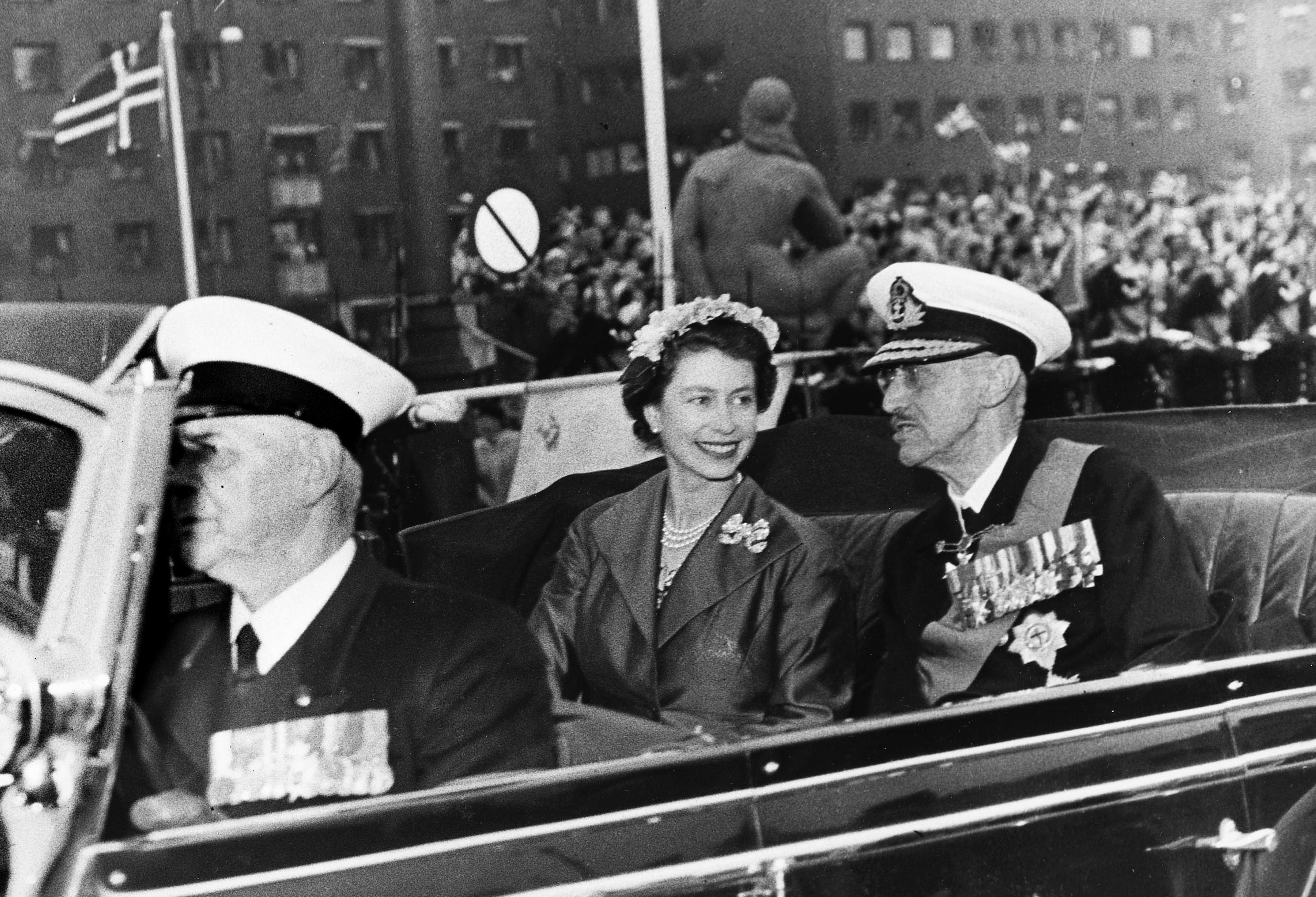 Sort-hvitt bilde av Kong Haakon og Dronning Elizabeth i åpen bil. Dronningen lytter til Kong Haakon med et stort smil.