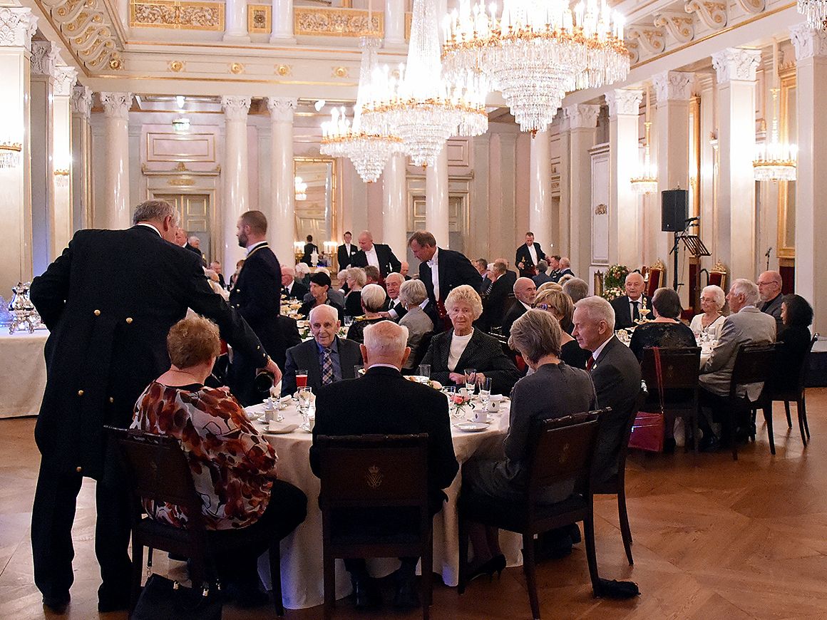 Gjester sitter rundt runde bord mens livrékledde fra hoffet sørger for serveringen.