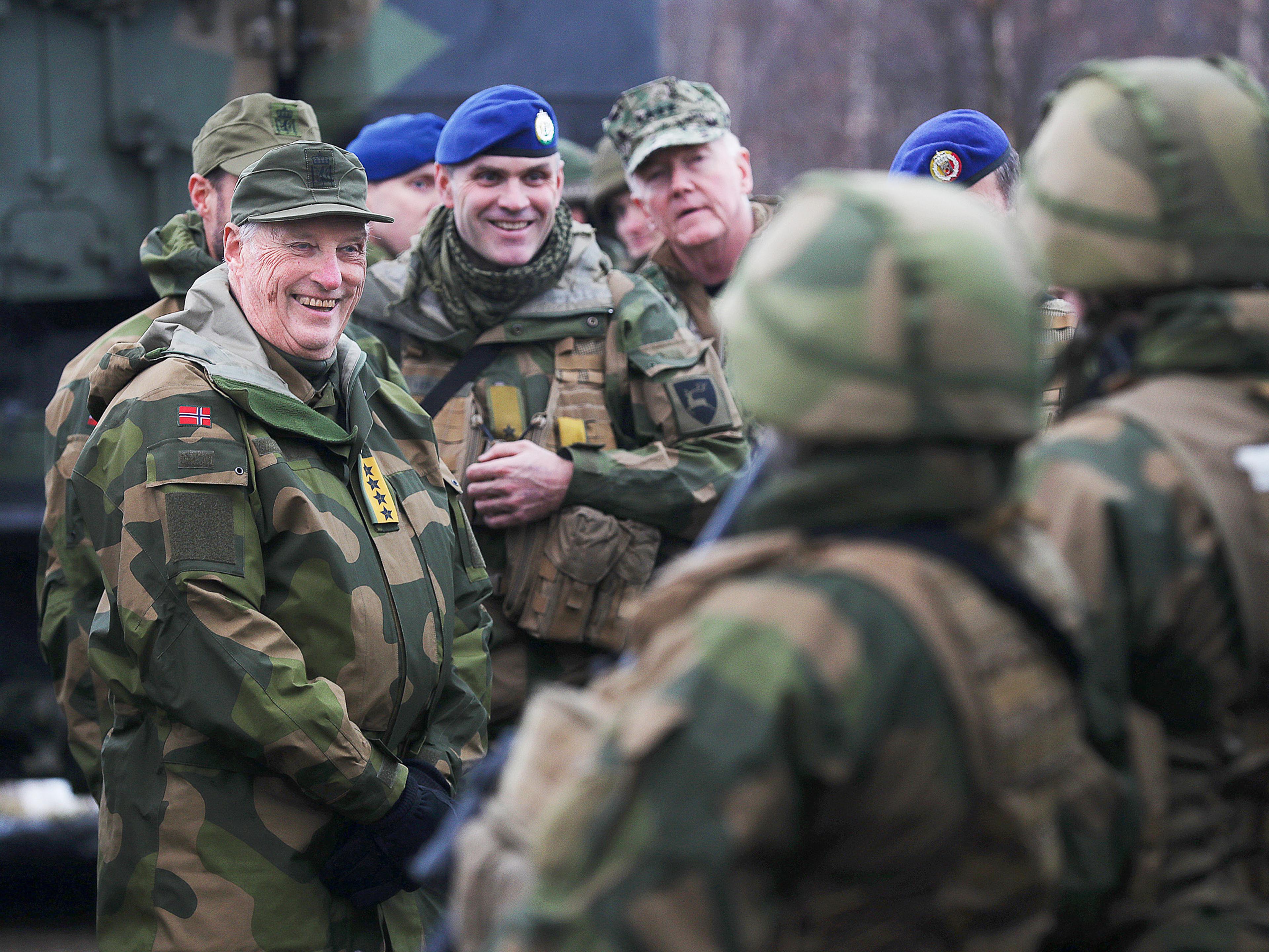Kong Harald kledd i Forsvarets membranuniform med Hærens feltlue smiler mot menige soldater.. Bak Kongen, ser man daværende brigadesjef og en amerikansk marineoffiser.