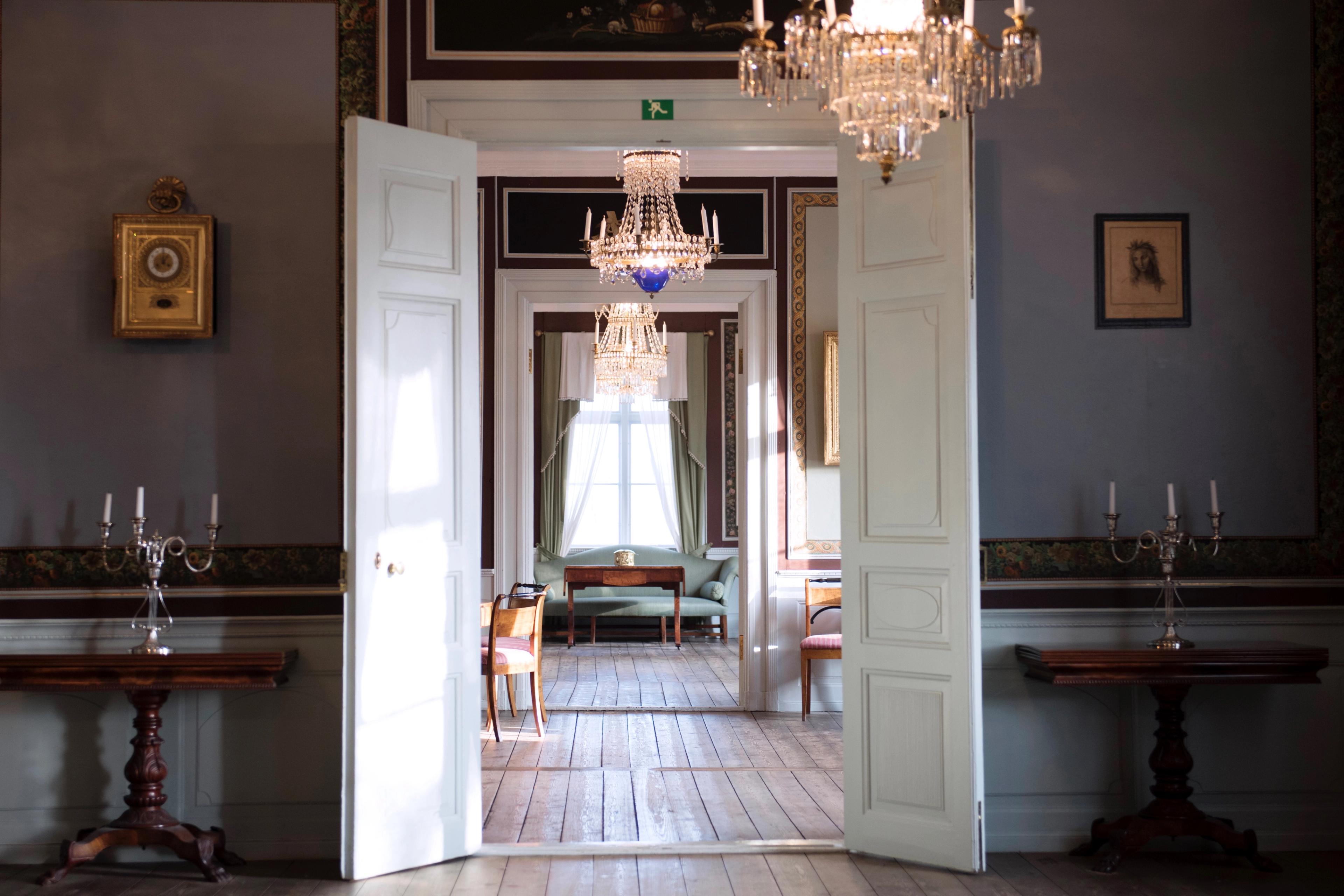 Store hvite dobbeltdører står åpne og vi kan se fra rom til rom innover. Det er tregulv og lysekroner i rommene.