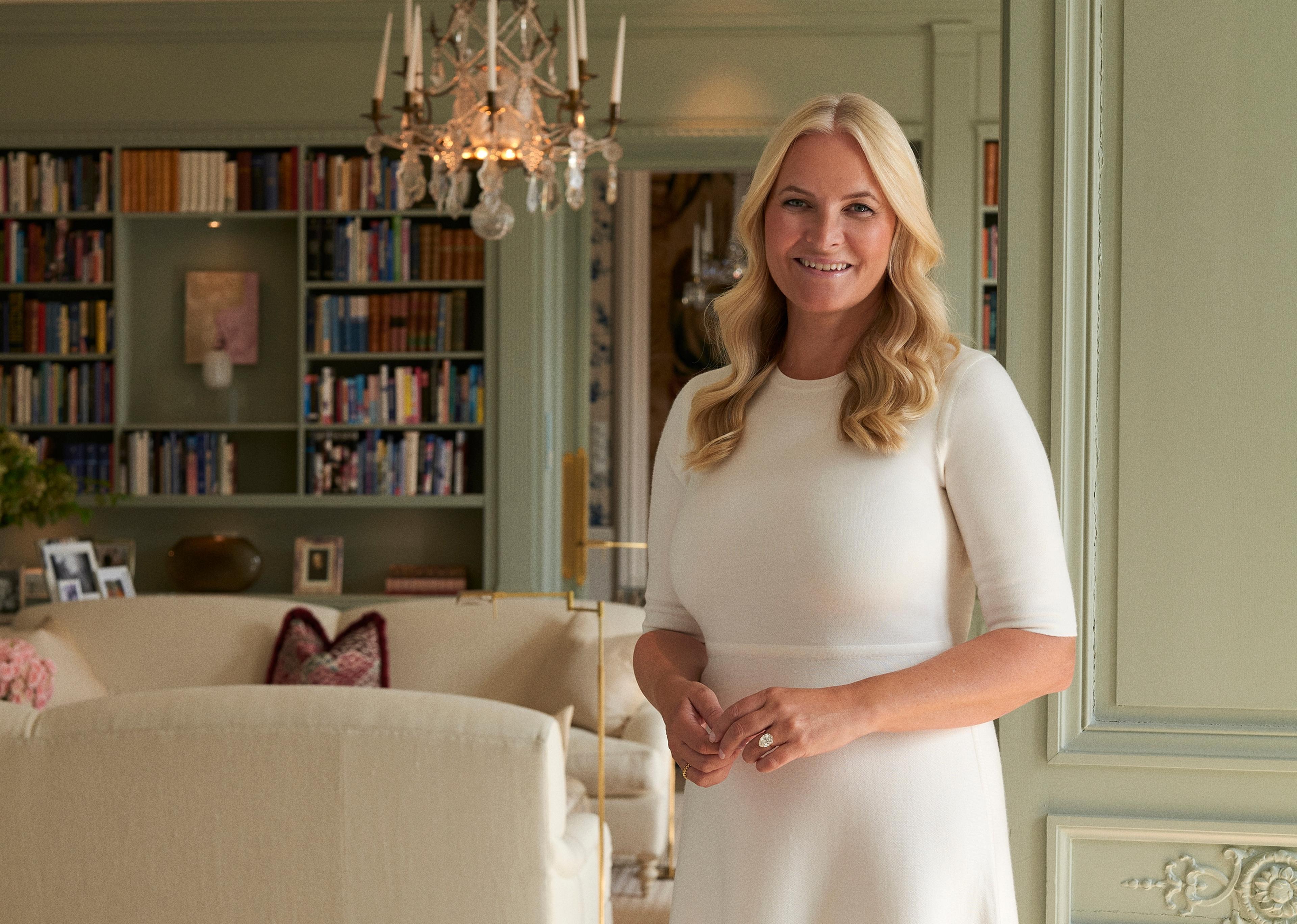 Kronprinsessen står smilende i hvit kjole i døren inn i en stue med mange bokhyller.