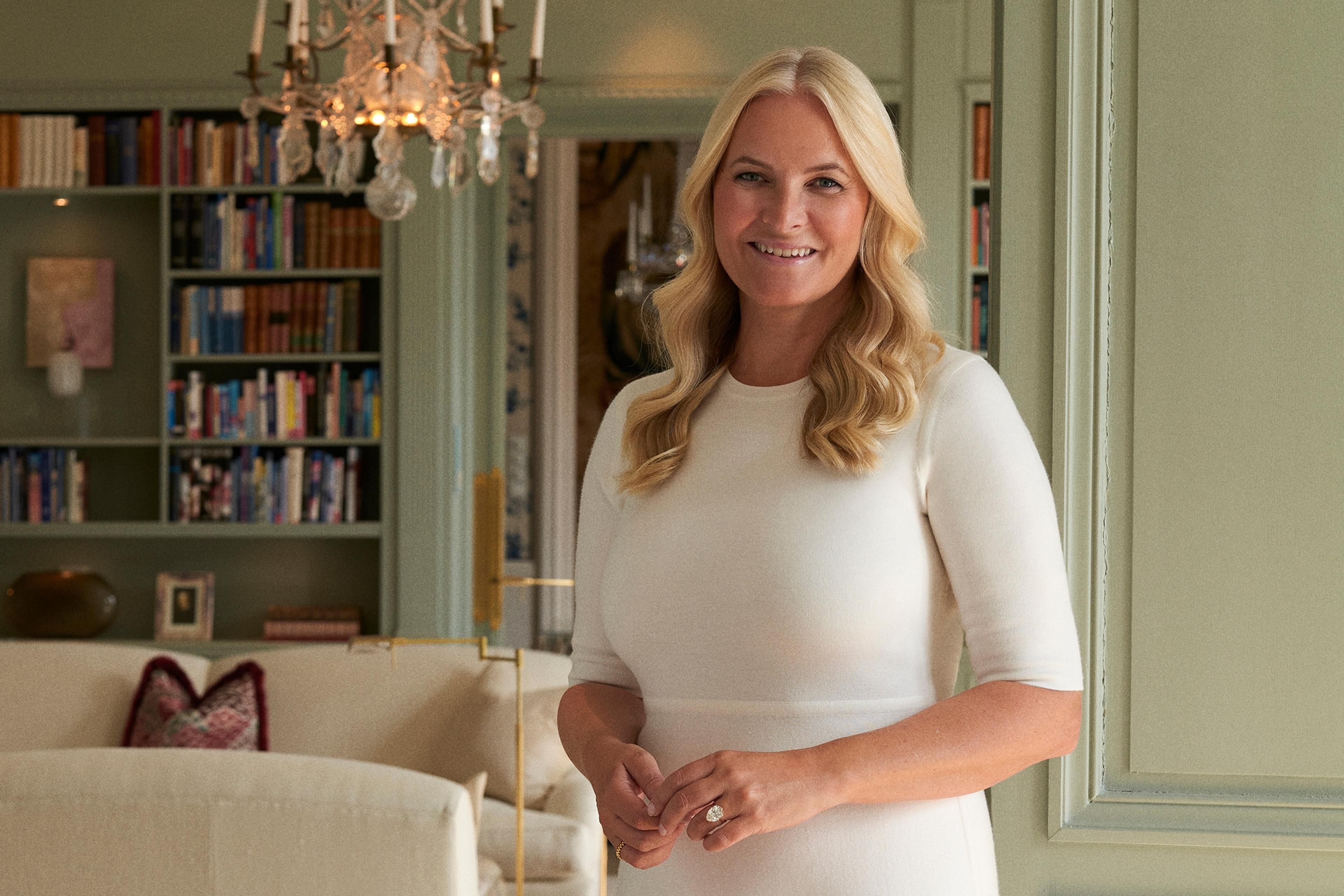 Kronprinsessen står smilende i hvit kjole i døren inn i en stue med mange bokhyller.