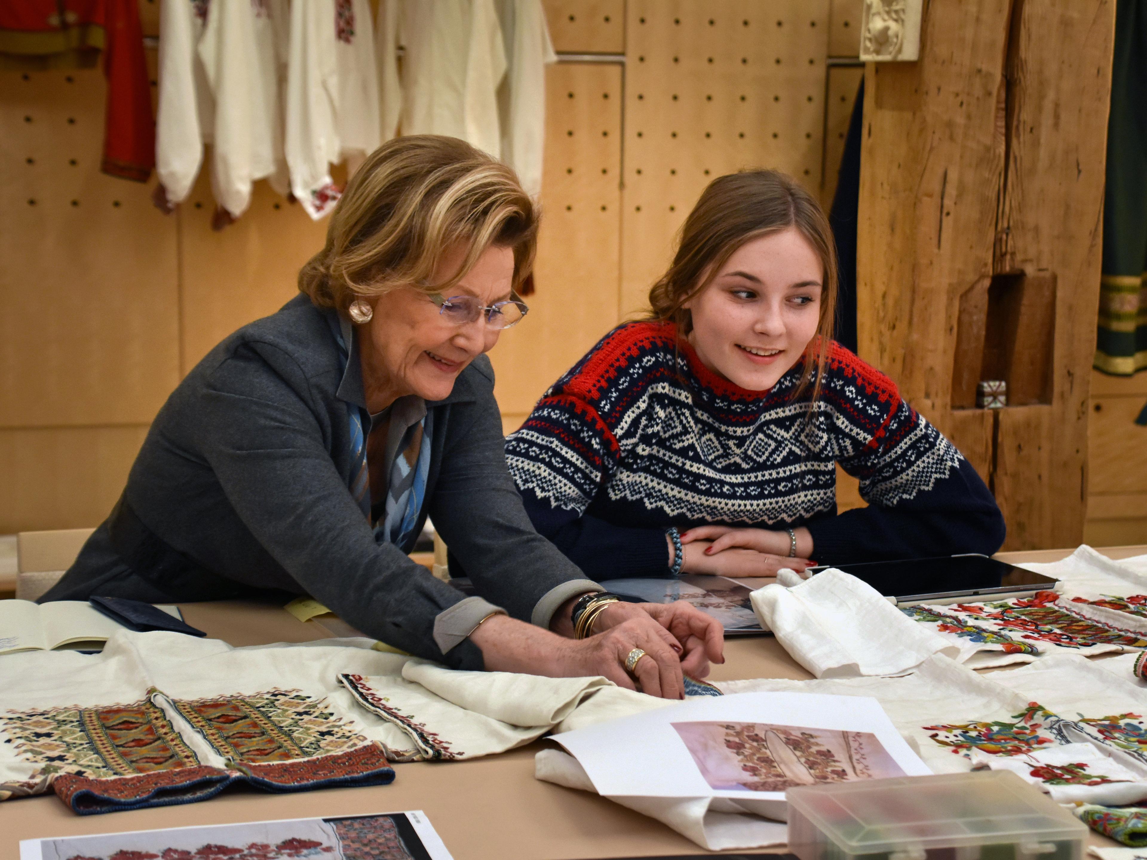 Dronning Sonja og Prinsesse Ingrid Alexandra sitter ved et arbeidsbord og ser på ulike stoff- og broderiprøver.