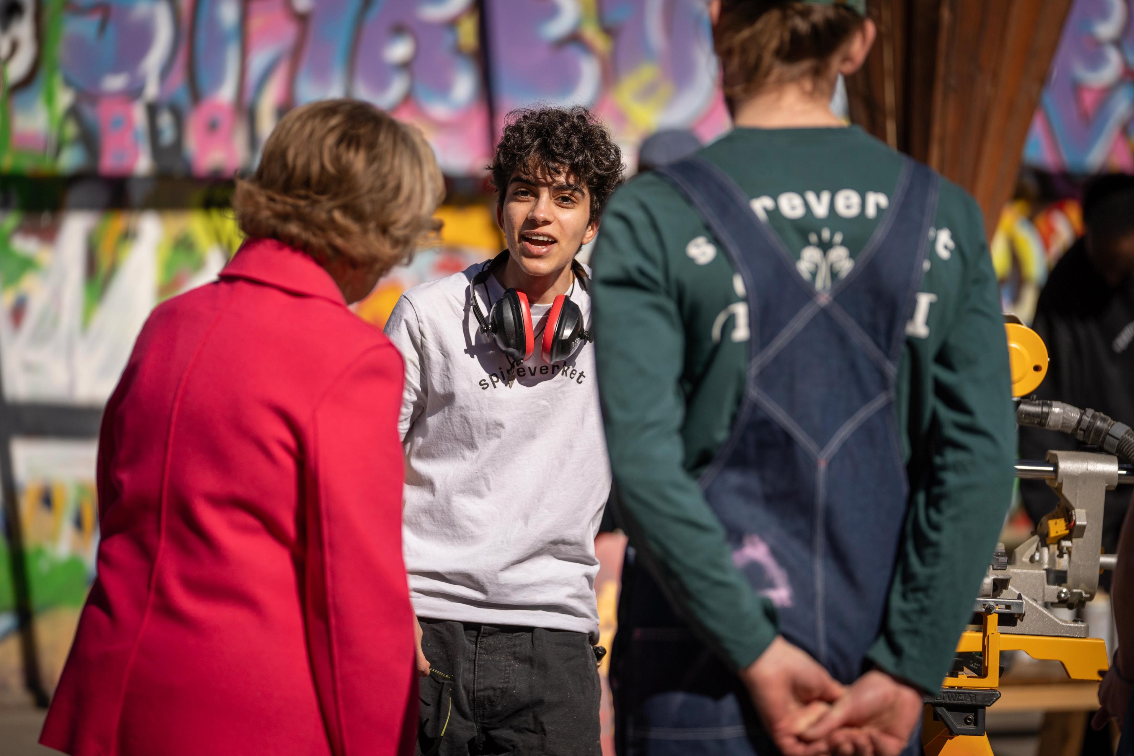 En ung gutt i samtale med Dronning Sonja med hørselsvern rundt halsen.