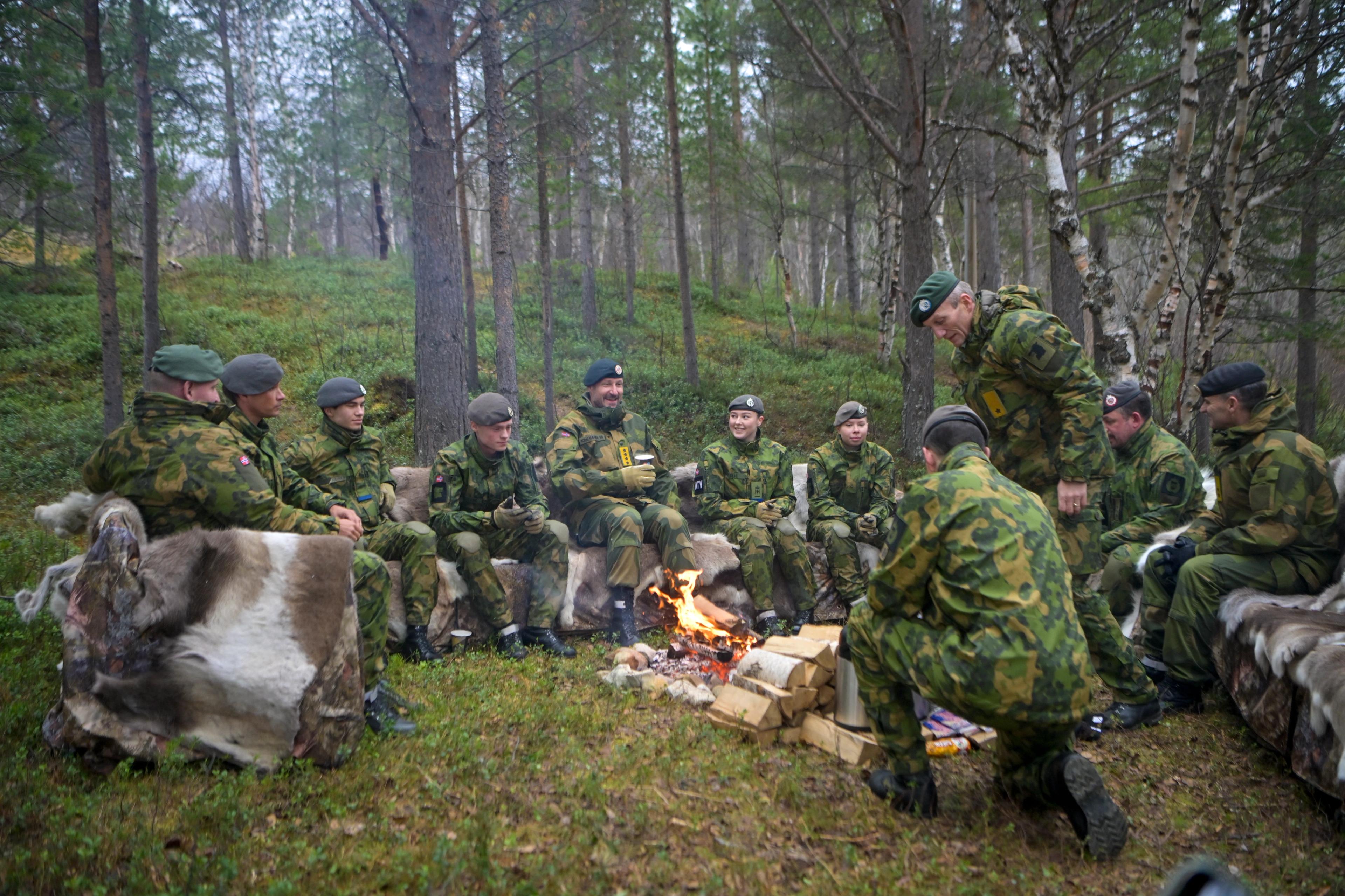 Kongen sitter på benker med reinskinn sammen med soldater, alle i feltuniform, rundt et bål.