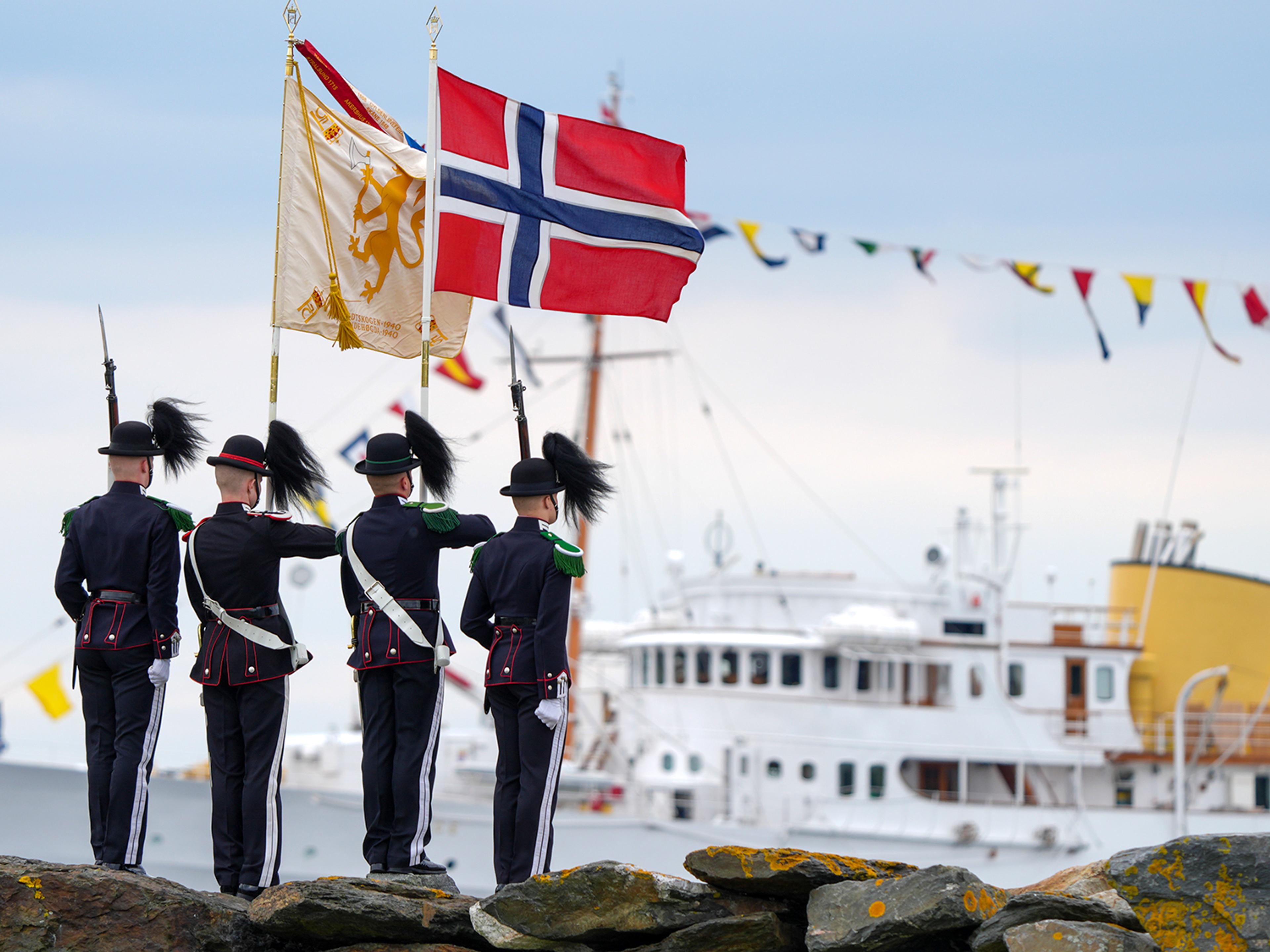 Vi ser ryggen til fire gardister som står øverst på en molo, med det norske flagget og et banner, og ansiktene er vendt mot havet der Kongeskipet Norge ligger ankret opp.