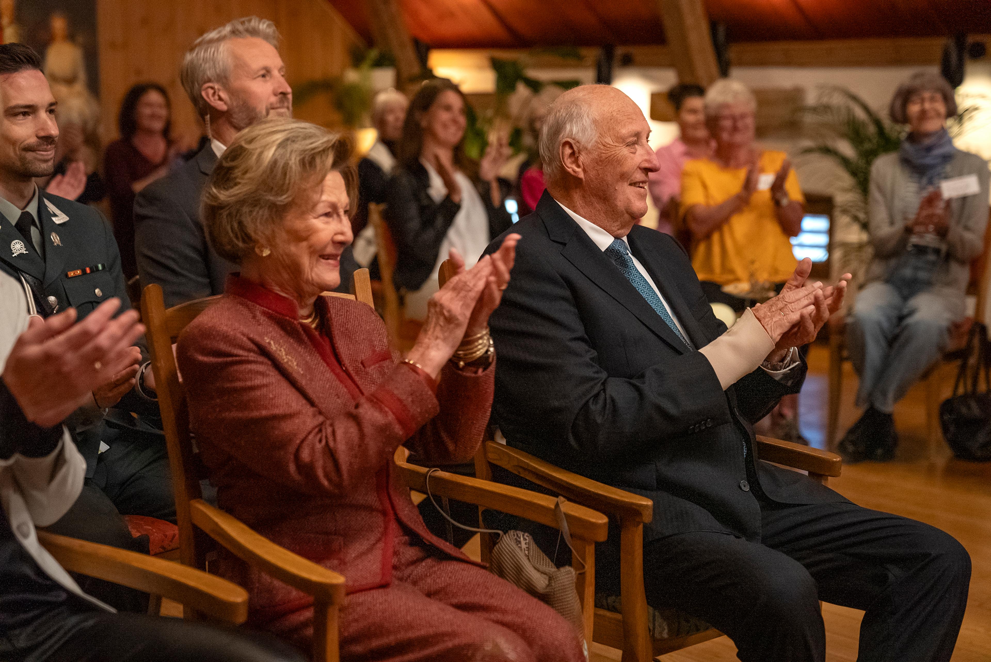 Kongen og Dronningen sitter omgitt av andre tilskuere og klapper spmilende etter en forestilling.