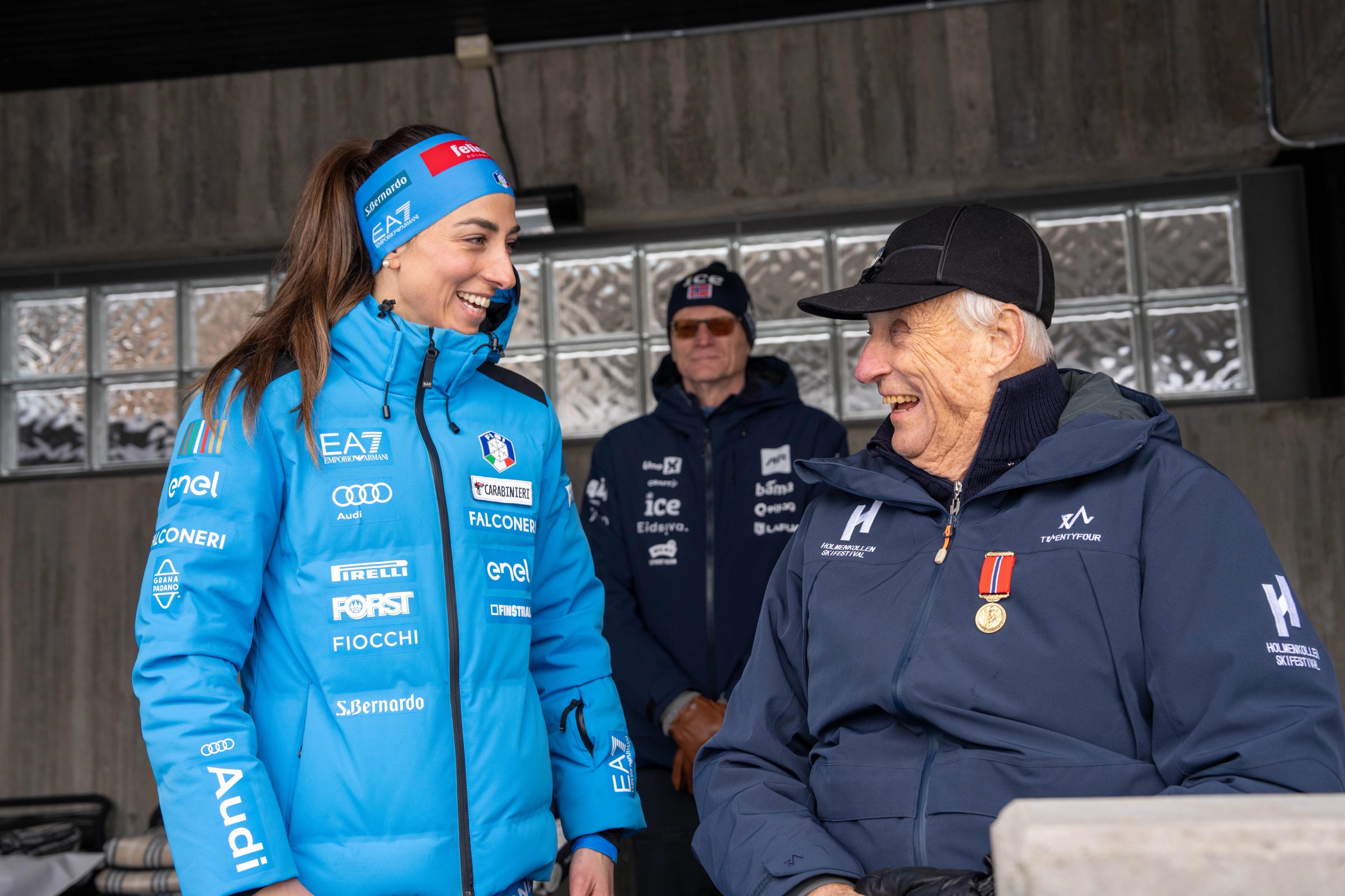 Skiskytteren Lisa Vittozzi fra Italia hilser på Kong Harald. De ser mot hverandre og ler. Kong Harald har Holmenkollmedaljen på brystet.