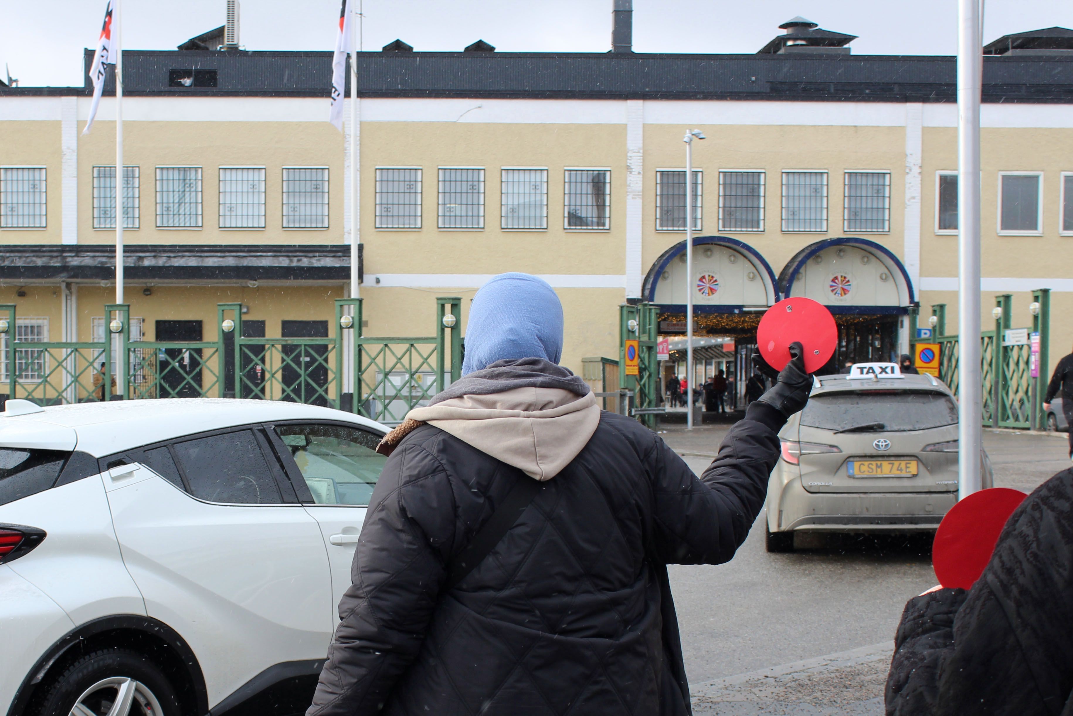 A person holds up a red paper circle in front of a road with cars and a yellow building in the background.