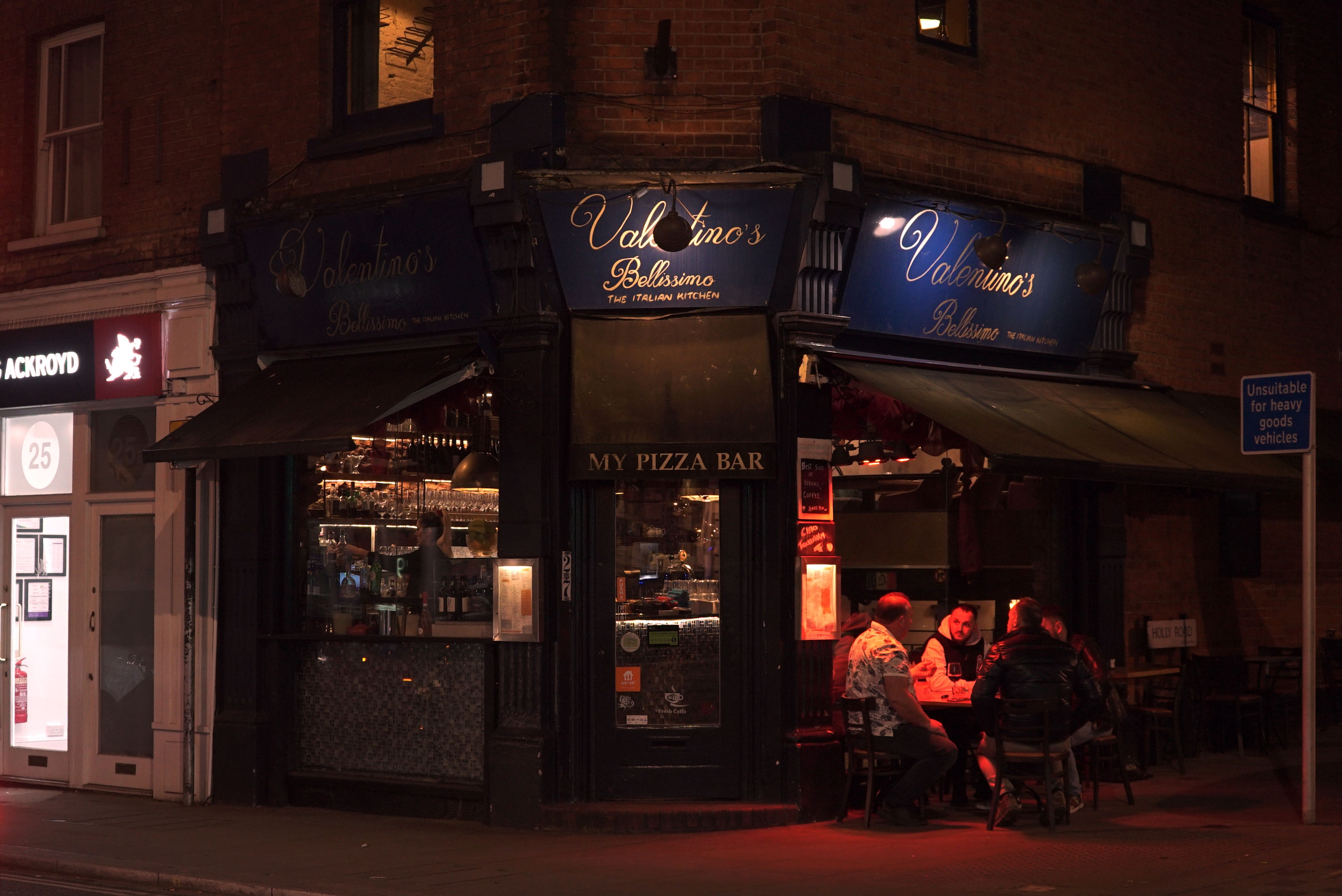 Photo of a pizza bar on a corner at night with people sat outside with heaters.