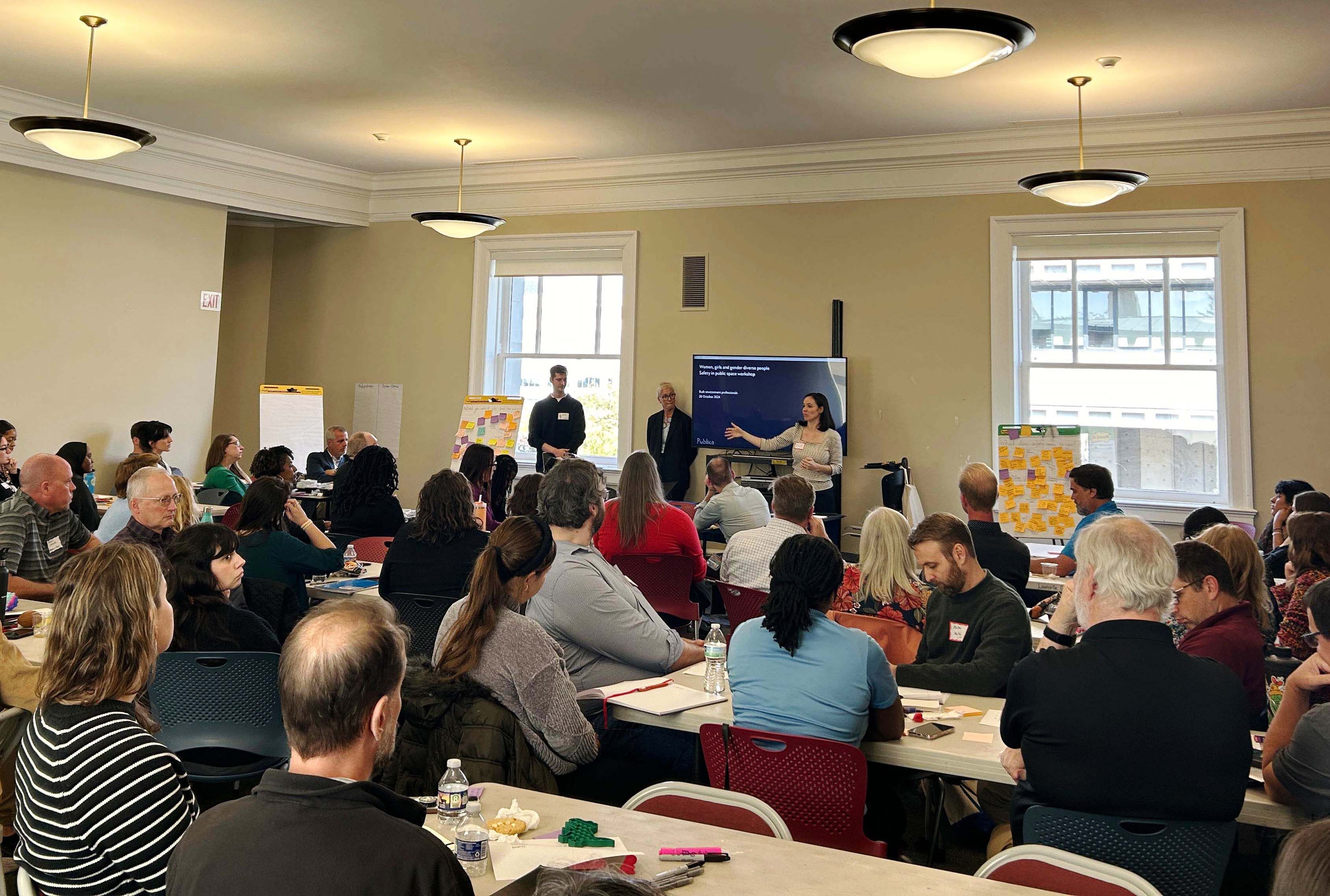 A room full of people sitting at desks facing a group of three people giving a presentation.