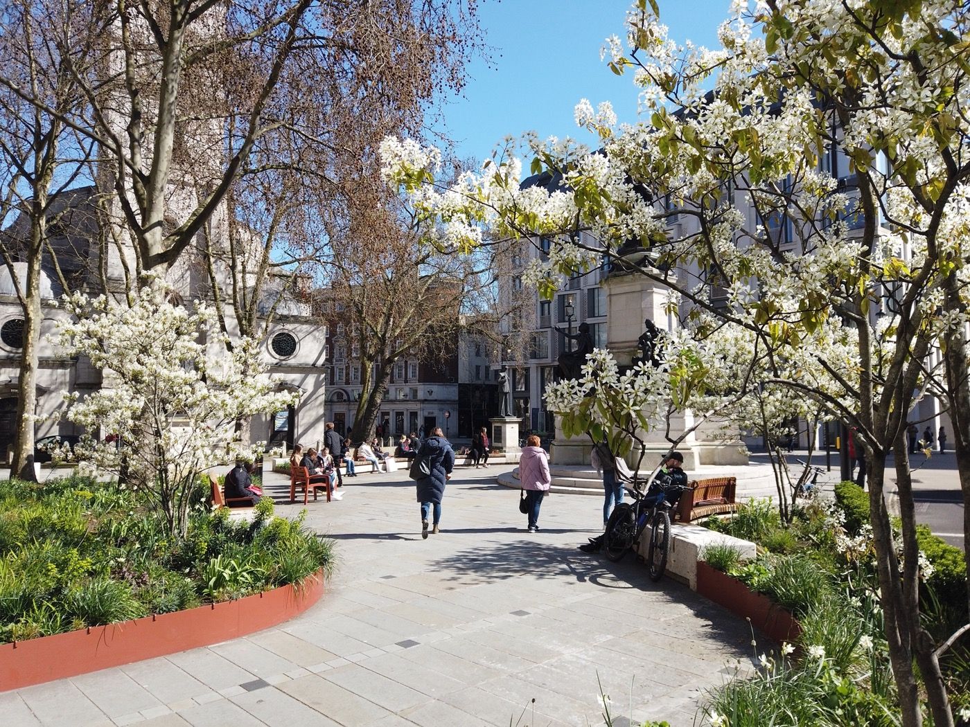 Photograph showing the pedestrianised public space at Strand Aldwych, featuring newly paved walking areas, modern wooden benches, orange metal planters, and blooming white trees in front of historic buildings.