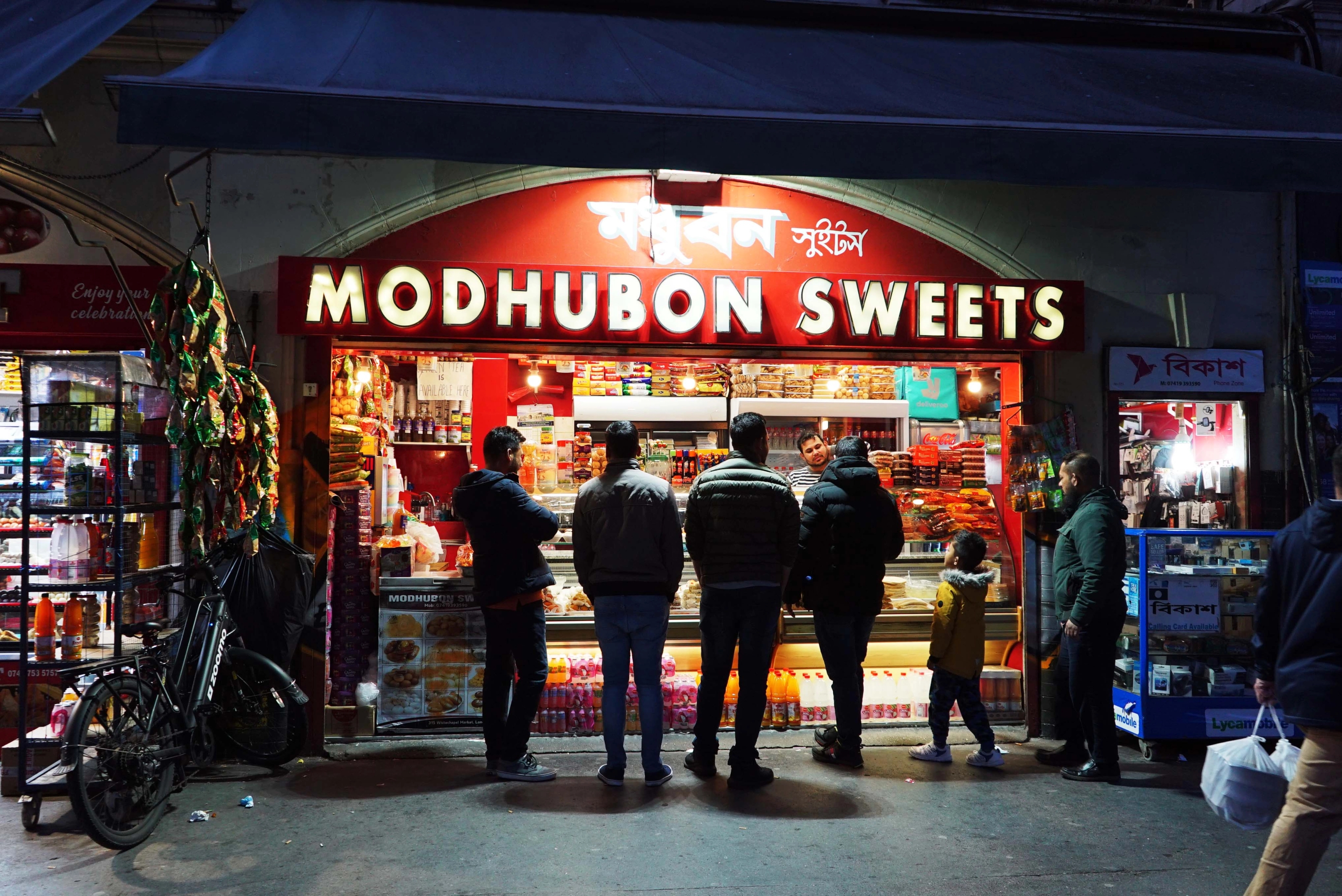 Photo of a lit shop front at night with a sign saying Modhubon sweets and people waiting out infront.