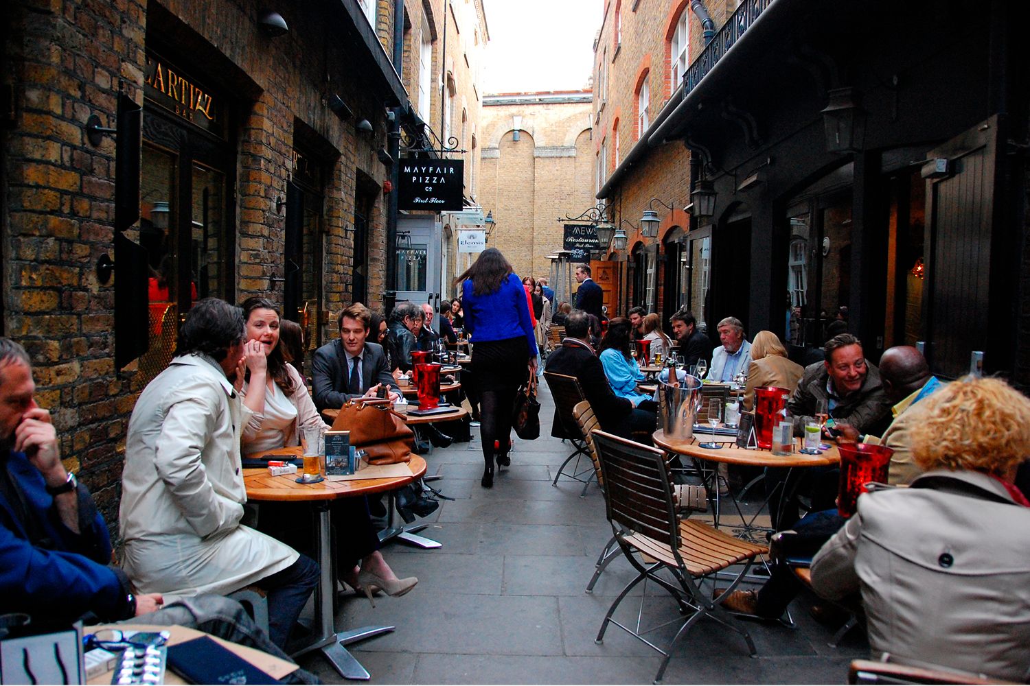 A pedestrian walks through the narrow space between outdoor tables and chairs with people sitting the length of the narrow street.
