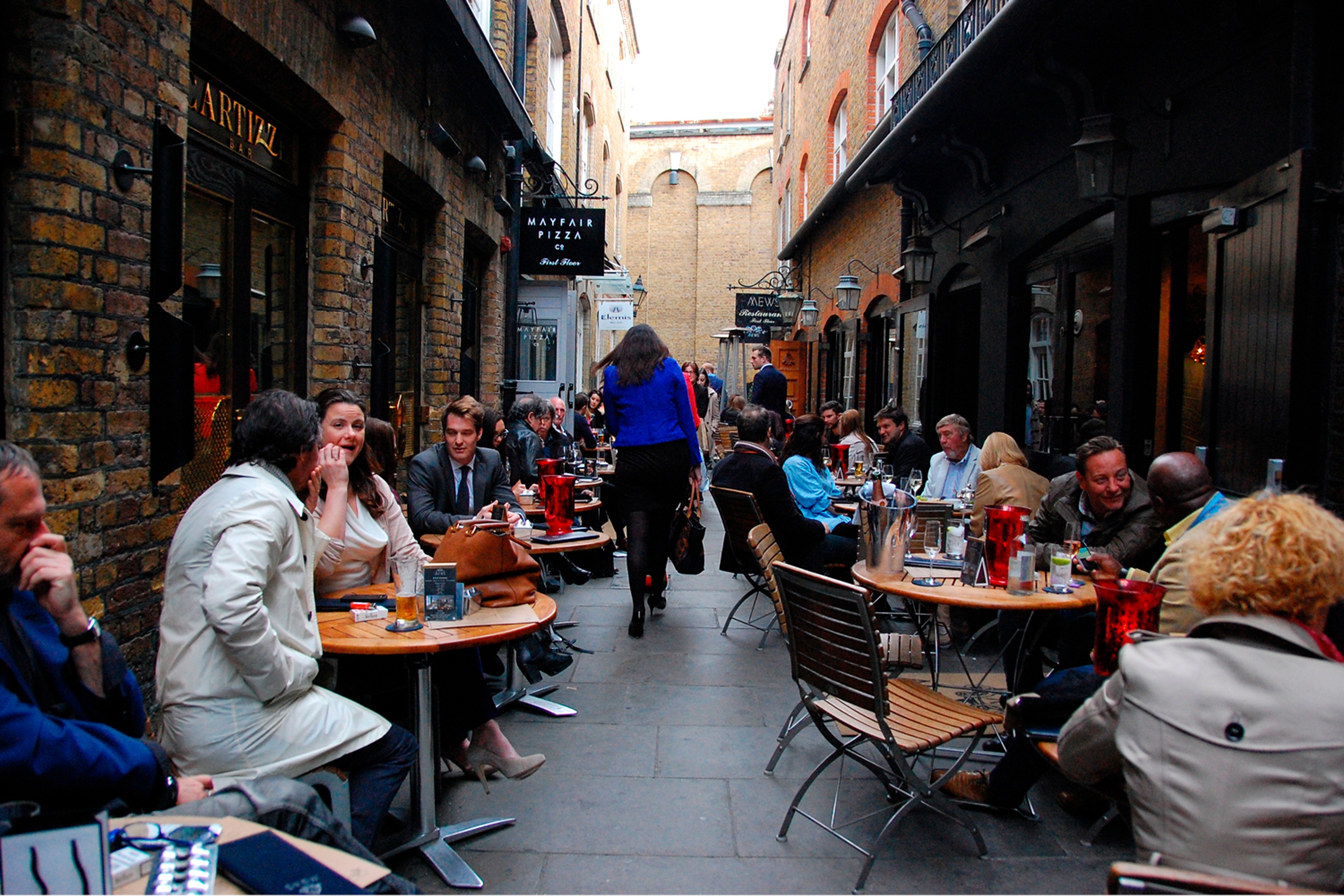 A pedestrian walks through the narrow space between outdoor tables and chairs with people sitting the length of the narrow street.