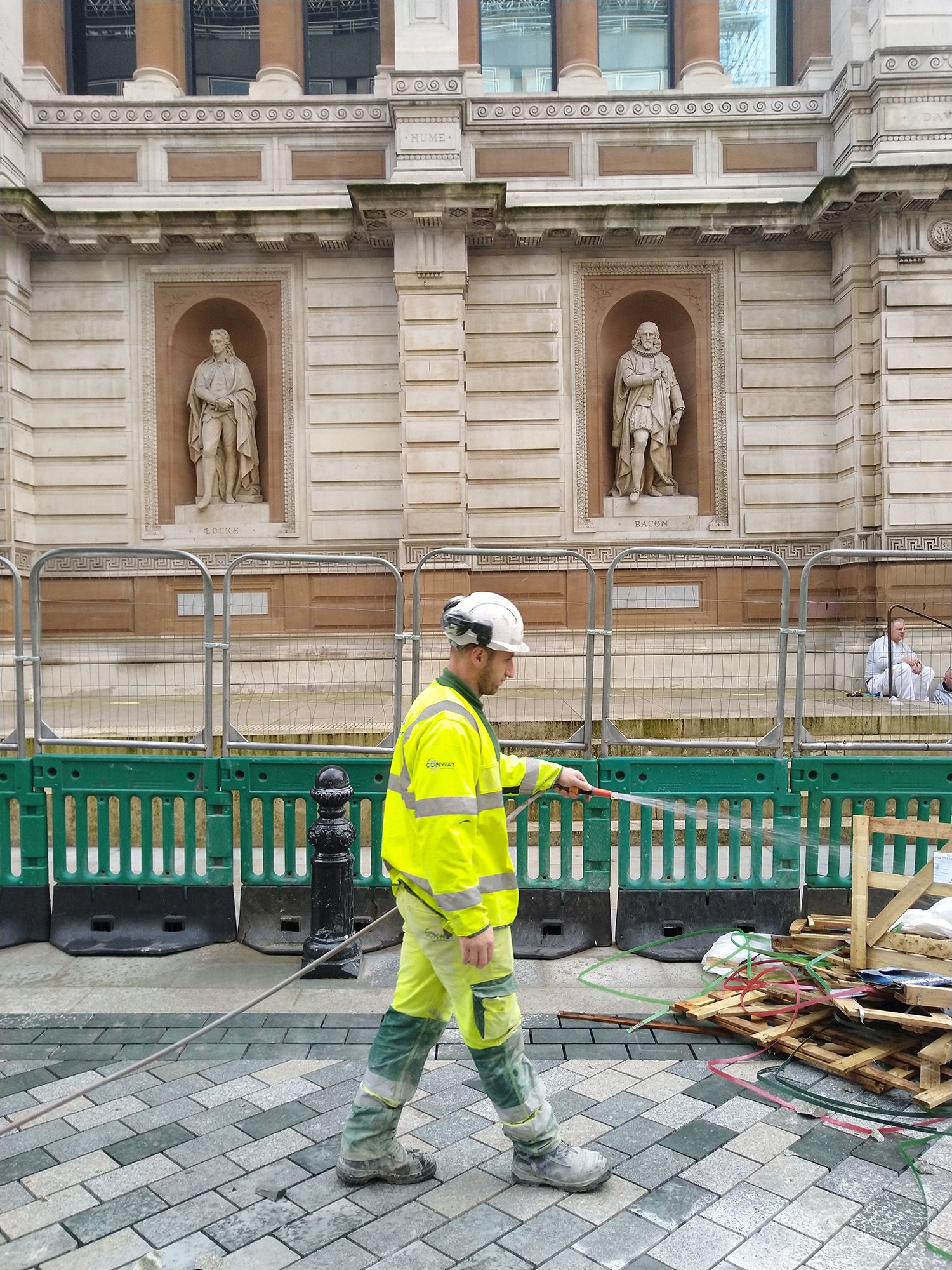 Construction worker wearing high-visibility hosing down a pile of wood in front of stone building with statues.