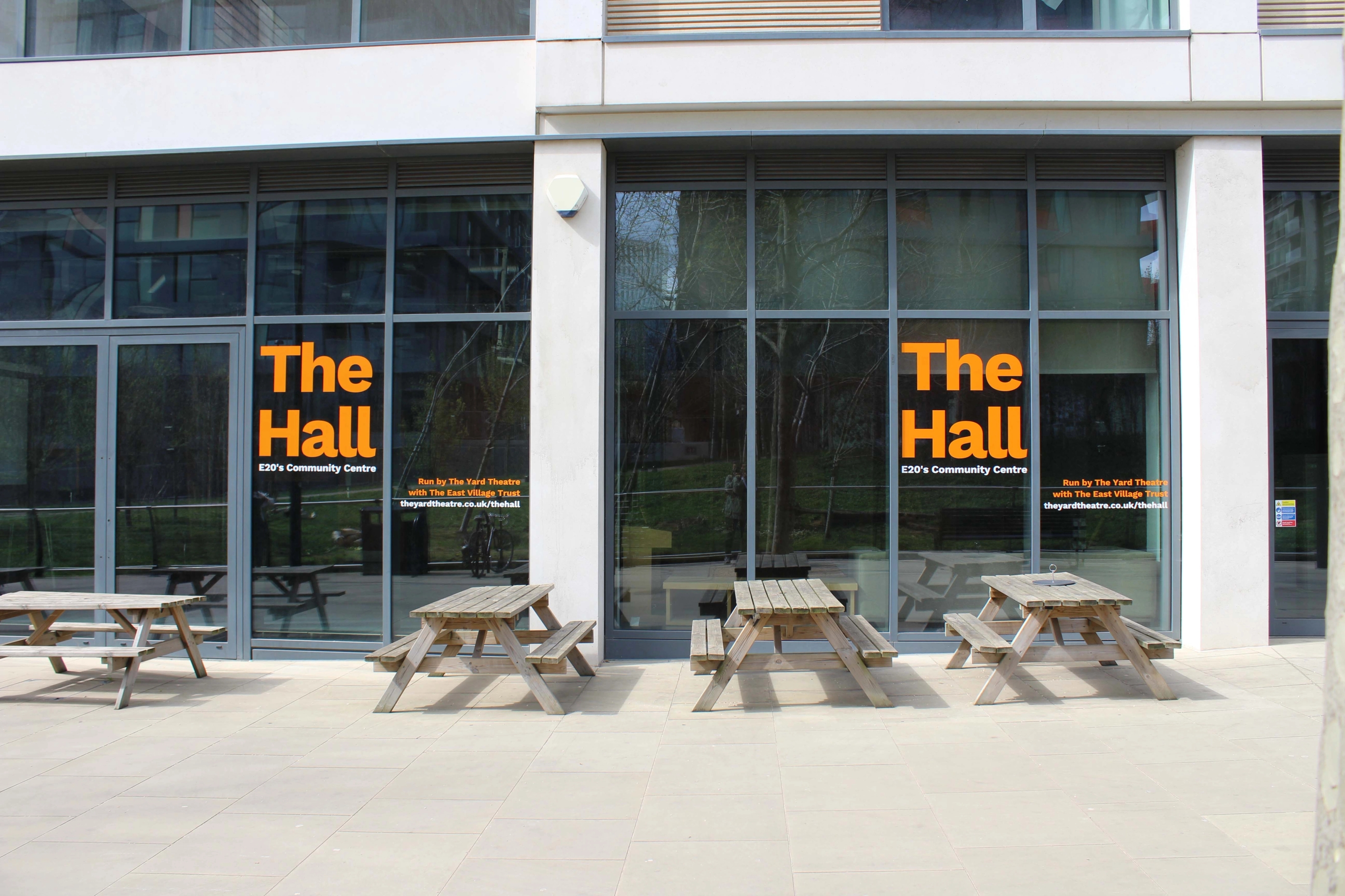 Wooden picnic tables outside a community space with large double height windows.