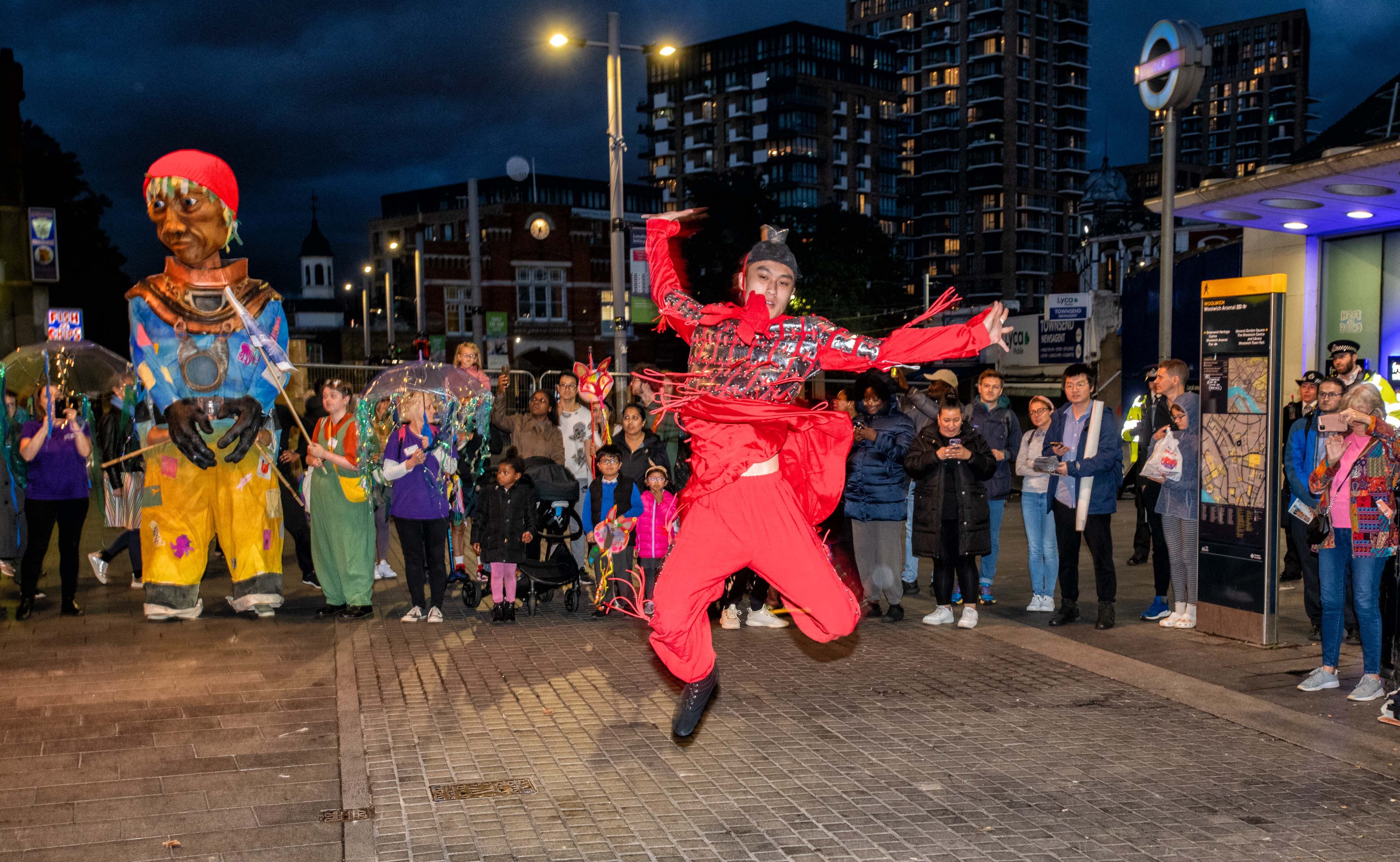 A performer dances in front of a crowd and a giant puppet outside Woolwich Arsenal DLR station at night.