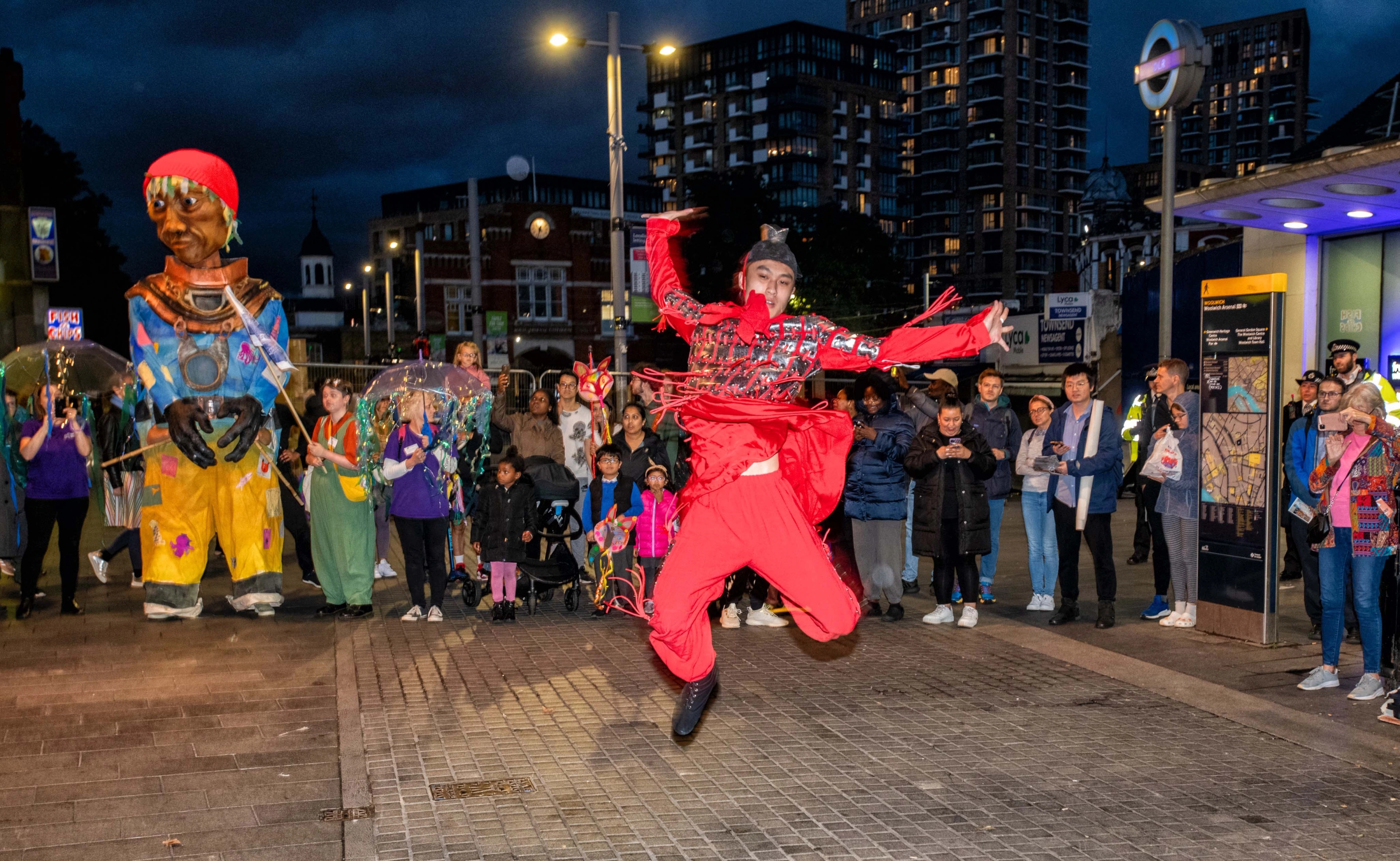 A performer dances in front of a crowd and a giant puppet outside Woolwich Arsenal DLR station at night.