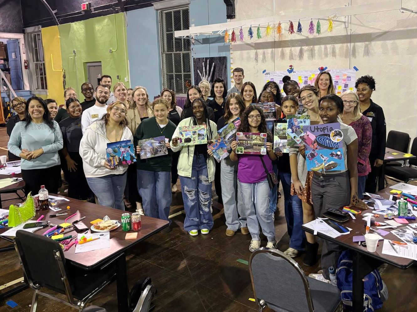A large group stands together with some people holding up collages in a community space.