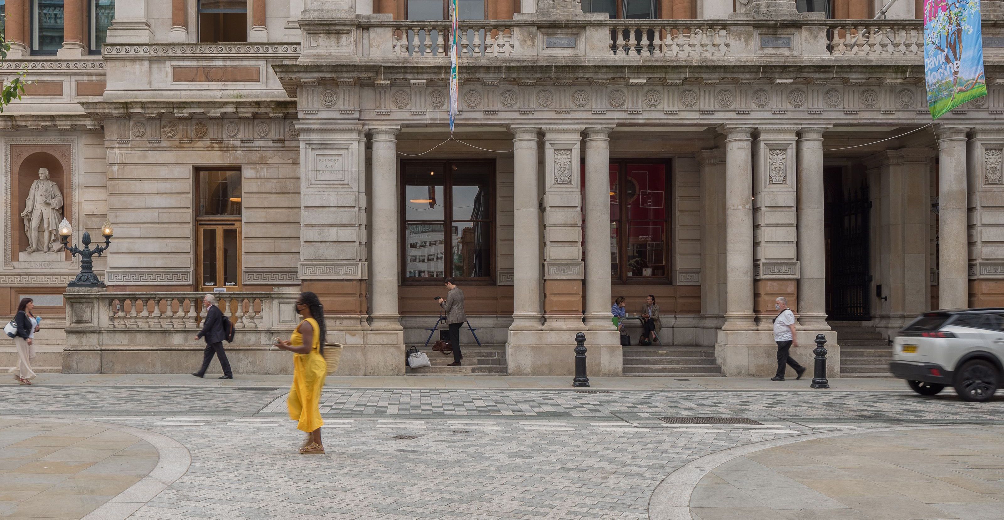 A pedestrian in a yellow dress crosses a flush carriageway in from of the Royal Academy.