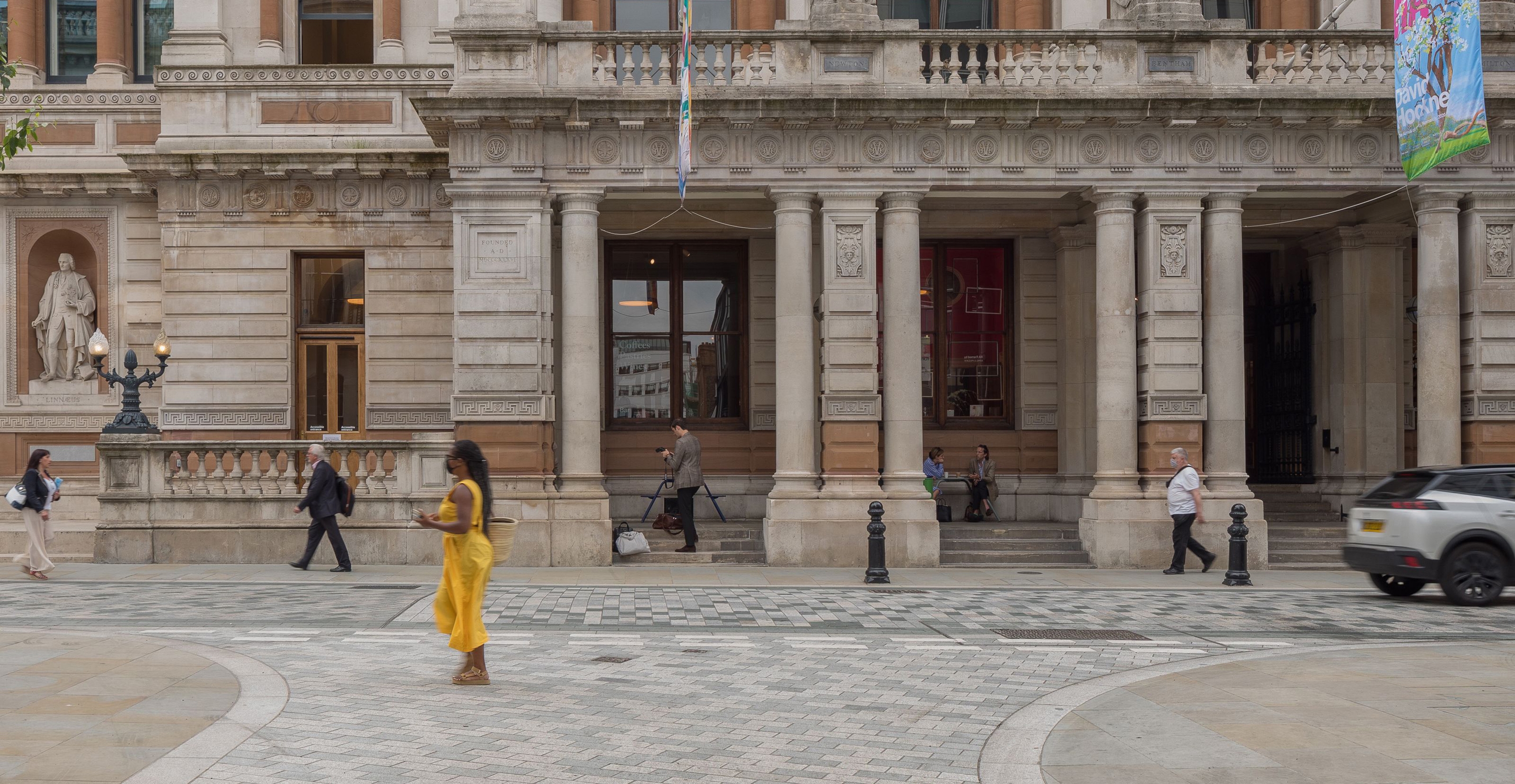 A pedestrian in a yellow dress crosses a flush carriageway in from of the Royal Academy.