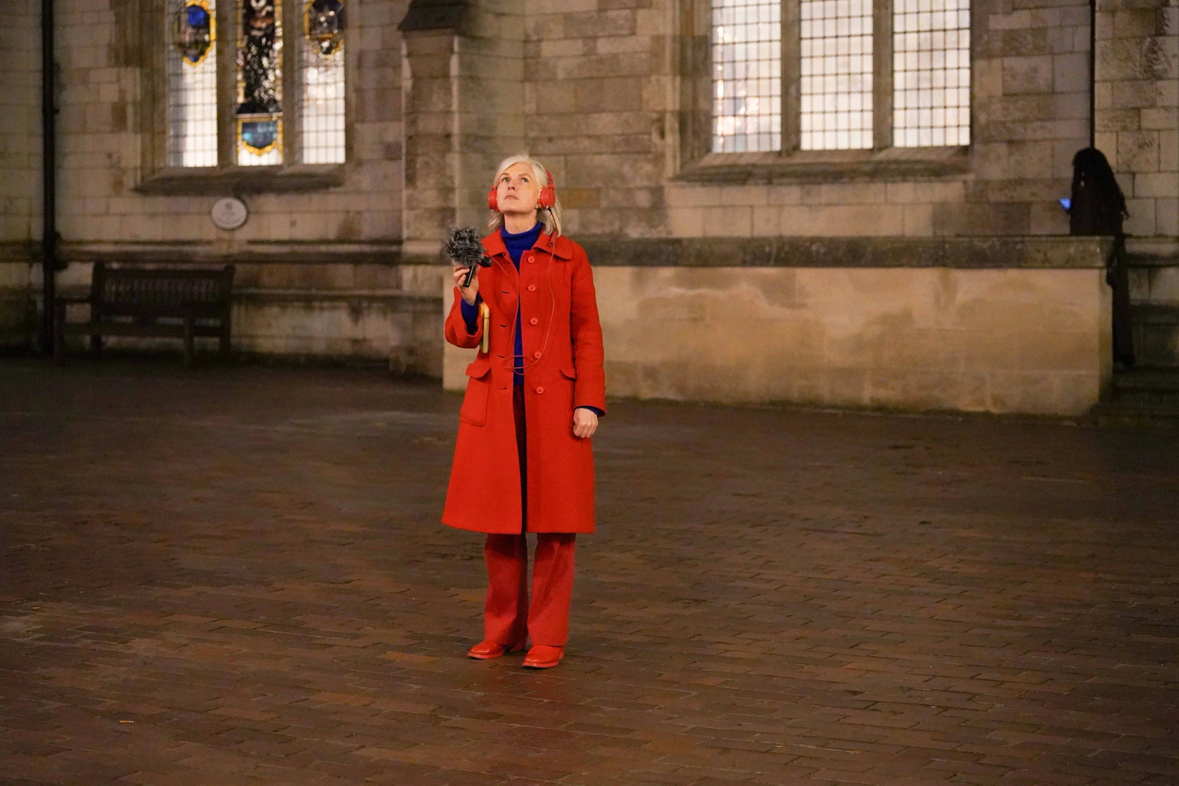 A person wearing a bright red jacket and boots looks up while holding up a microphone and wearing red headphones.