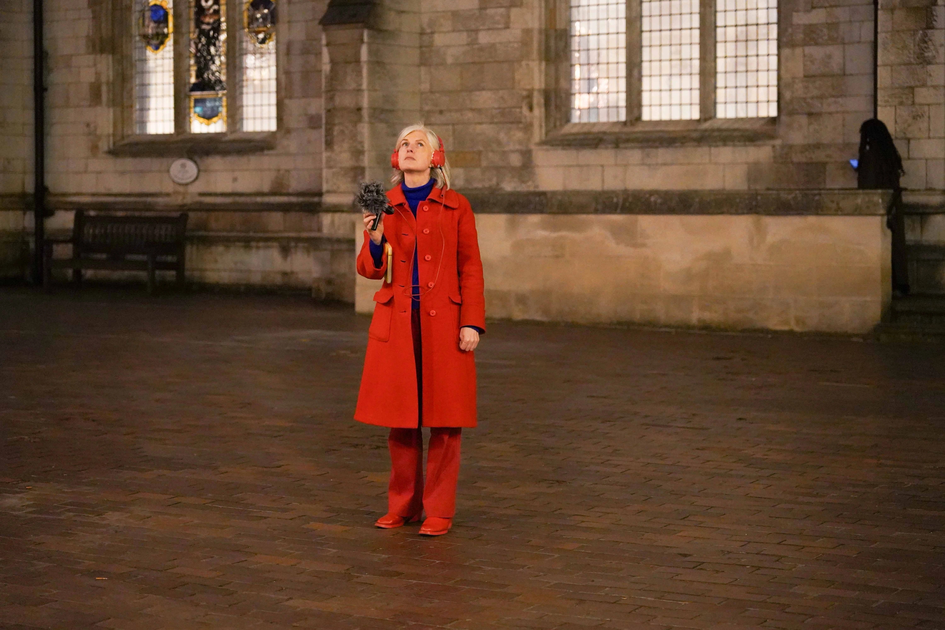 A person wearing a bright red jacket and boots looks up while holding up a microphone and wearing red headphones.